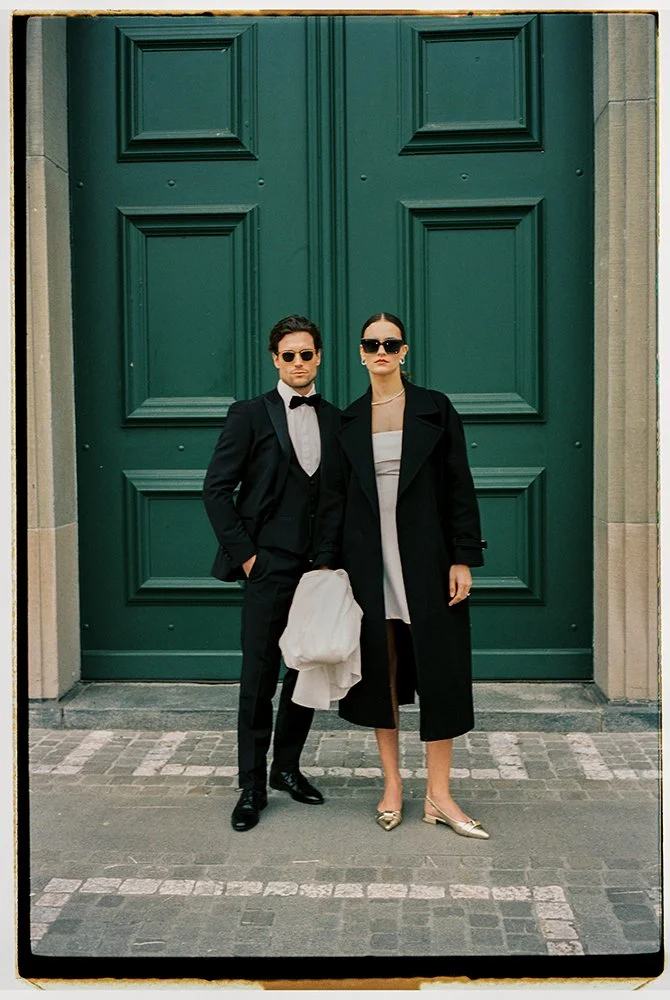 Ein Paar steht im Anzug und Mantel und Sonnenbrillen vor einer grossen grünen Türe.