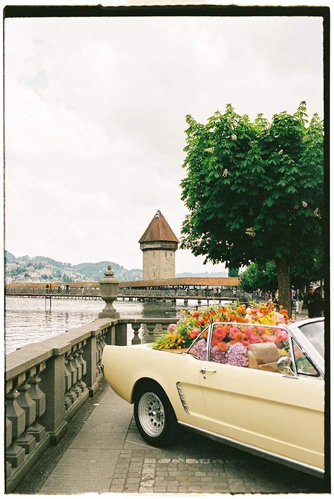 Ein hellgelber Oldtimer steht gefüllt mit bunten Blumen Vor der Kappelbrücke in Luzern.