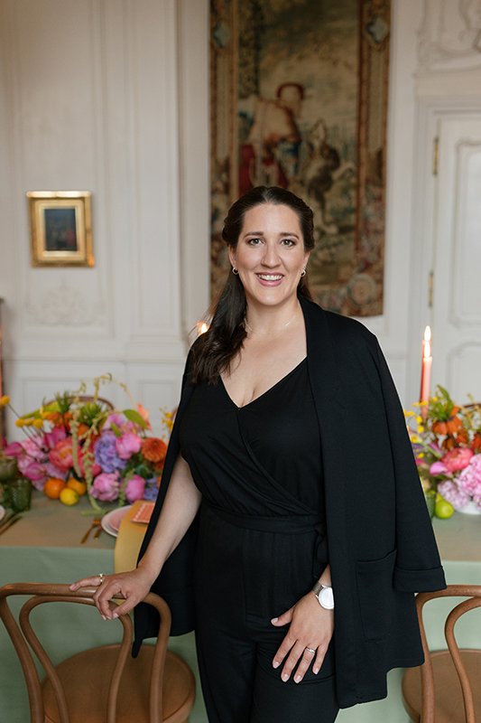 Eine Frau steht lachend in einem schwarzen Overall vor einem Tisch mit bunten Blumen.
