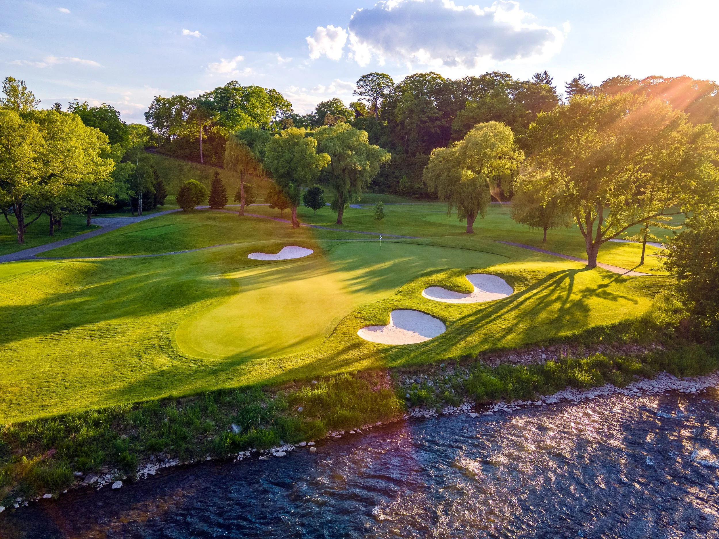 A scenic view of a golf course with green grass, sand bunkers, and a flagstick, surrounded by trees, with the sun shining and a partly cloudy sky overhead, next to a small creek.
