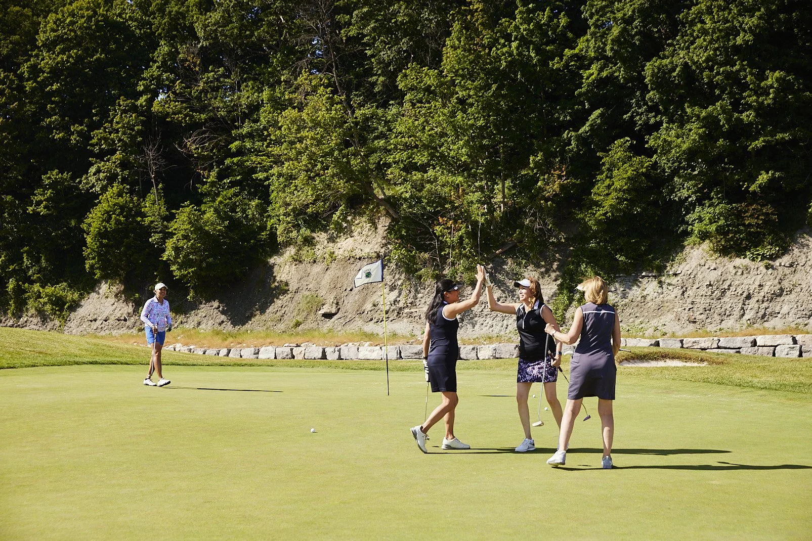 Four women celebrating on a golf green, with one woman in the background near a flagstick, all dressed in golf attire surrounded by trees and rocks.