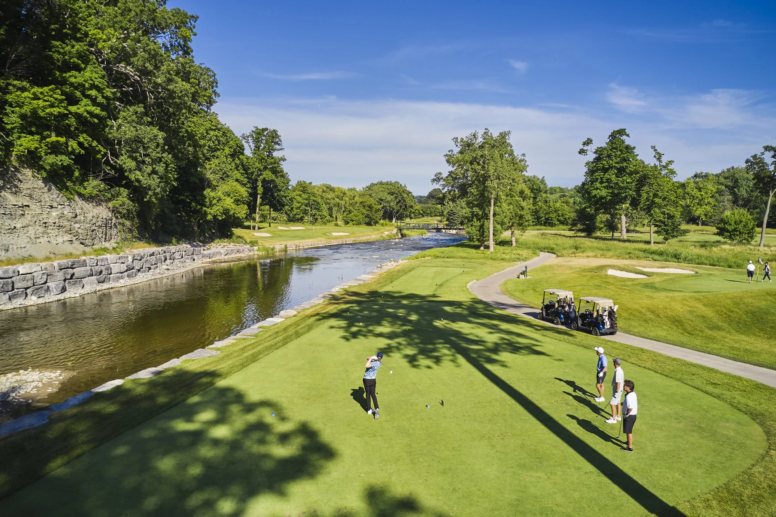 A golf course with people playing near a water hazard and golf carts parked nearby, surrounded by trees and clear blue sky.