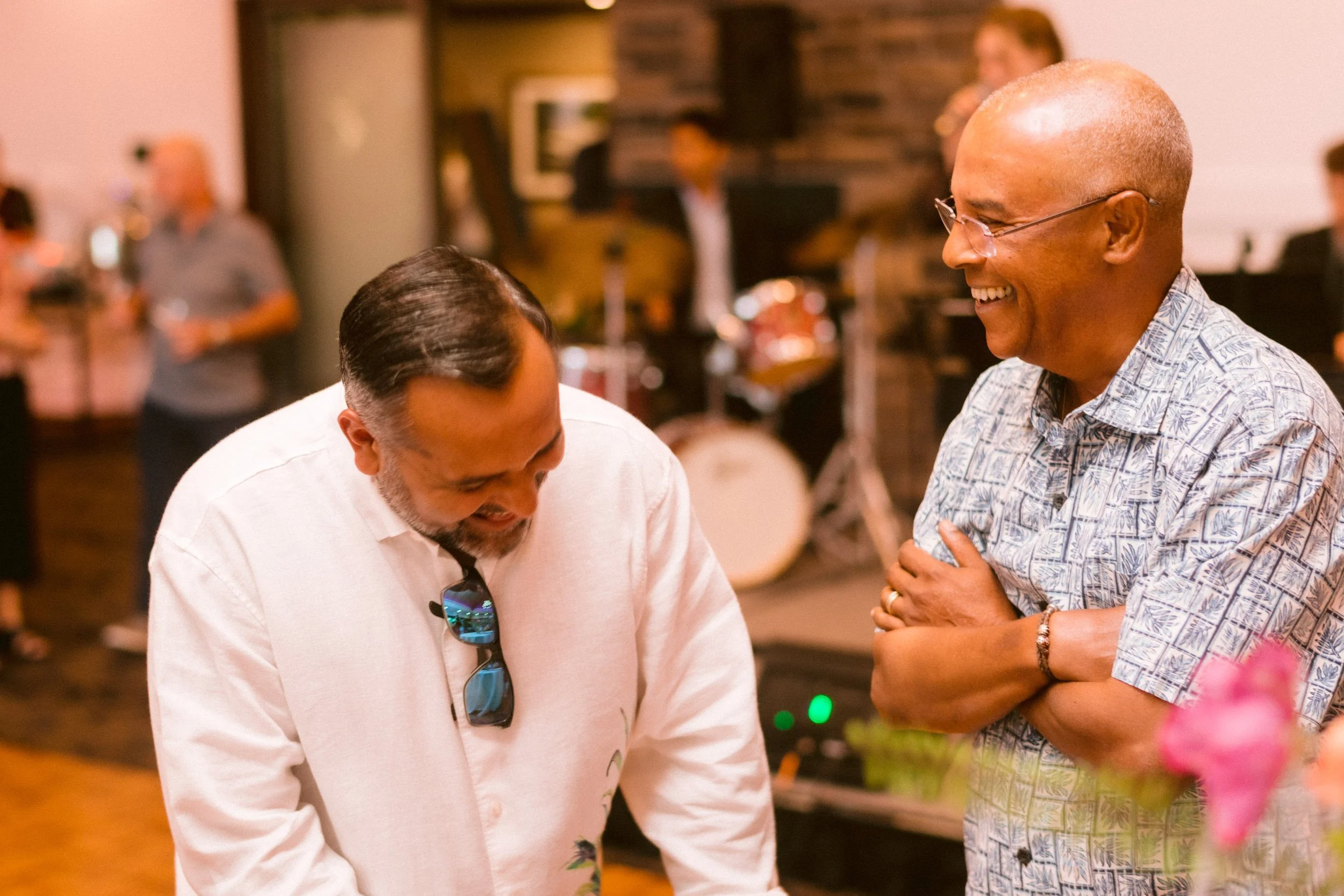 Two men laughing and talking in a social setting. One is wearing a white shirt with sunglasses hanging from the collar, the other is wearing glasses and a patterned shirt. Background shows other people and a brick wall.