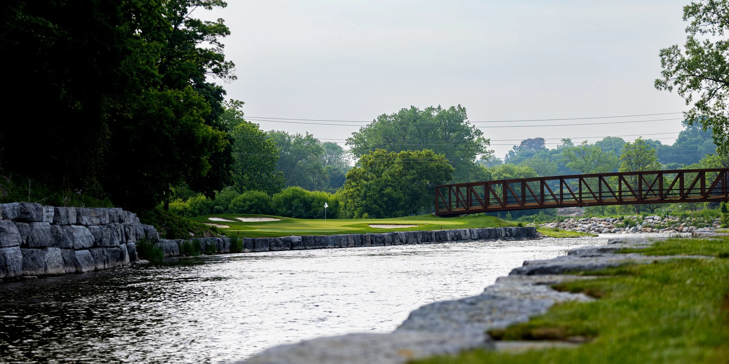 A scenic view of a golf course with a green, a flag, a small bridge over the Credit River, and trees surrounding the area.