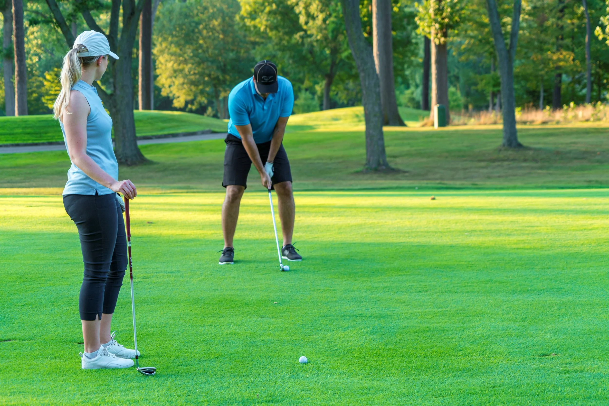 A young woman and man on a golf course, preparing to putt a golf ball. The woman is holding a putter and watching, while the man is bent over, aiming his putt. The setting is lush with green grass and trees.