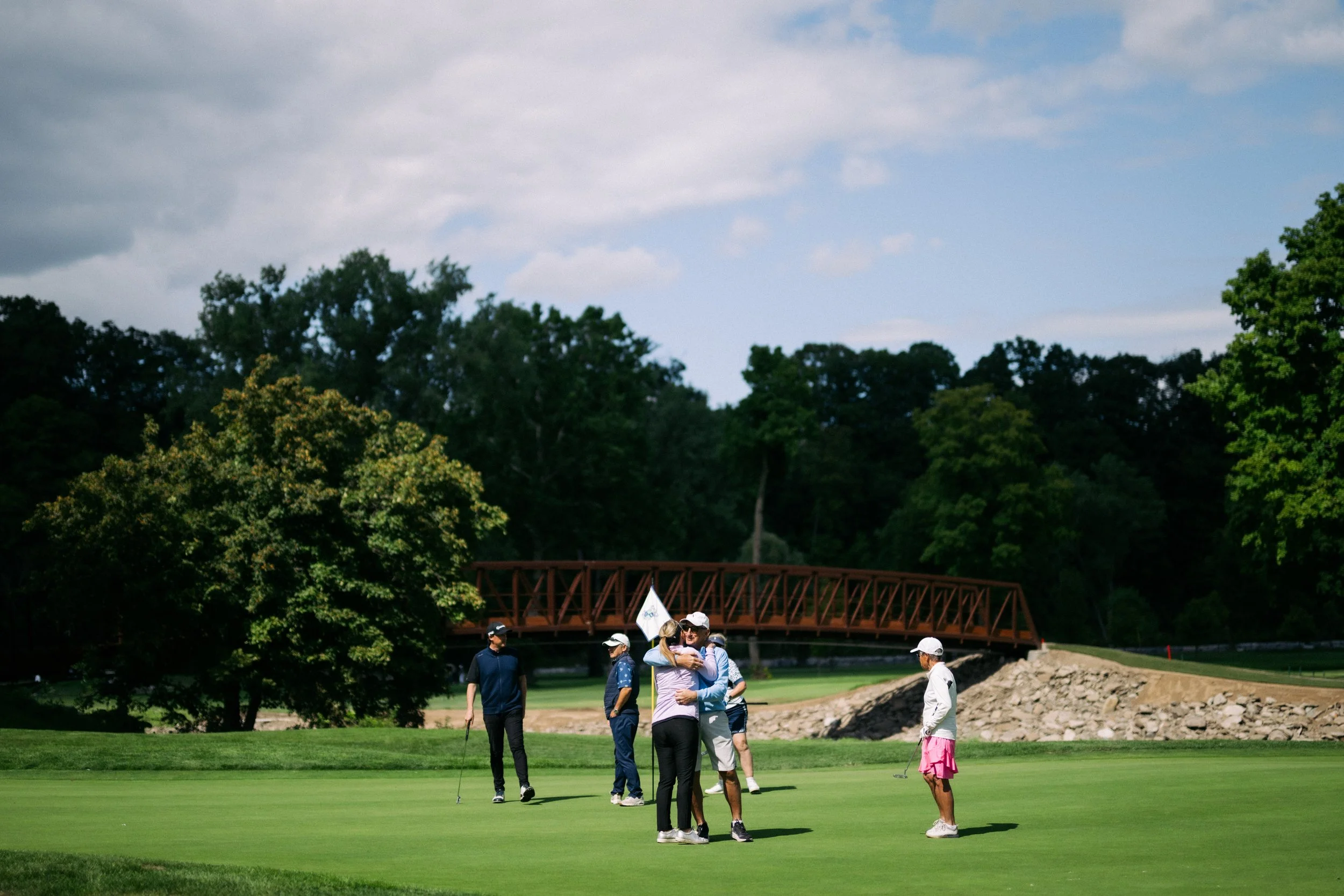 Group of people playing golf on a green course with trees, a bridge, and a cloudy sky in the background.