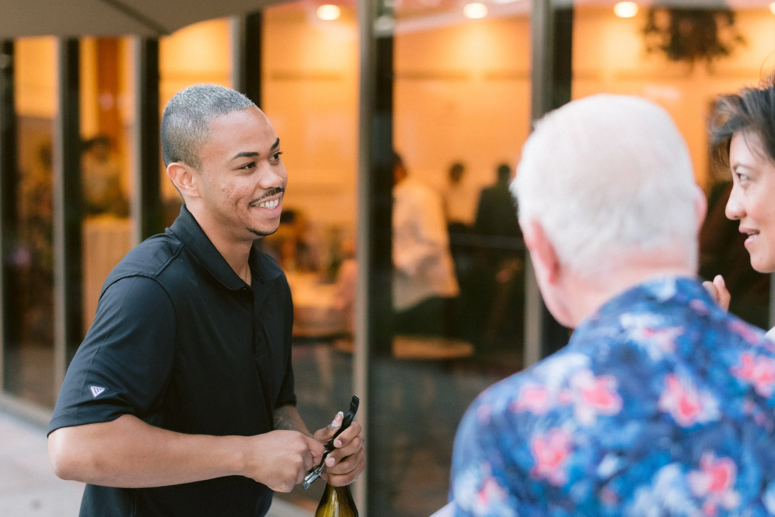 A man with short gray hair and a black polo shirt smiling and holding a bottle and opener, talking to a man with short white hair and another woman with dark brown hair, in front of a building with warm lighting.