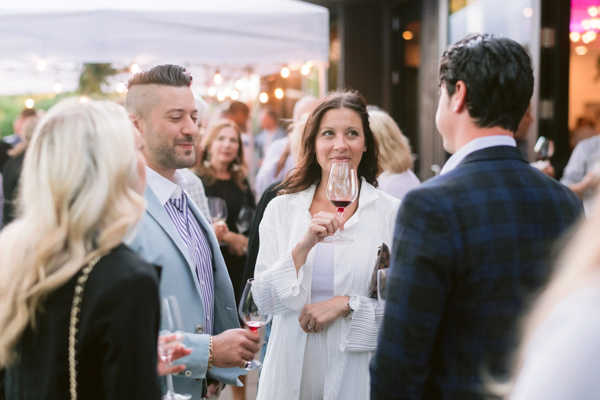 People dressed in formal attire socializing at an outdoor event during the evening. A woman in a white dress is holding a glass of red wine and is engaged in conversation with a man in a dark jacket. String lights are hanging overhead, creating a war