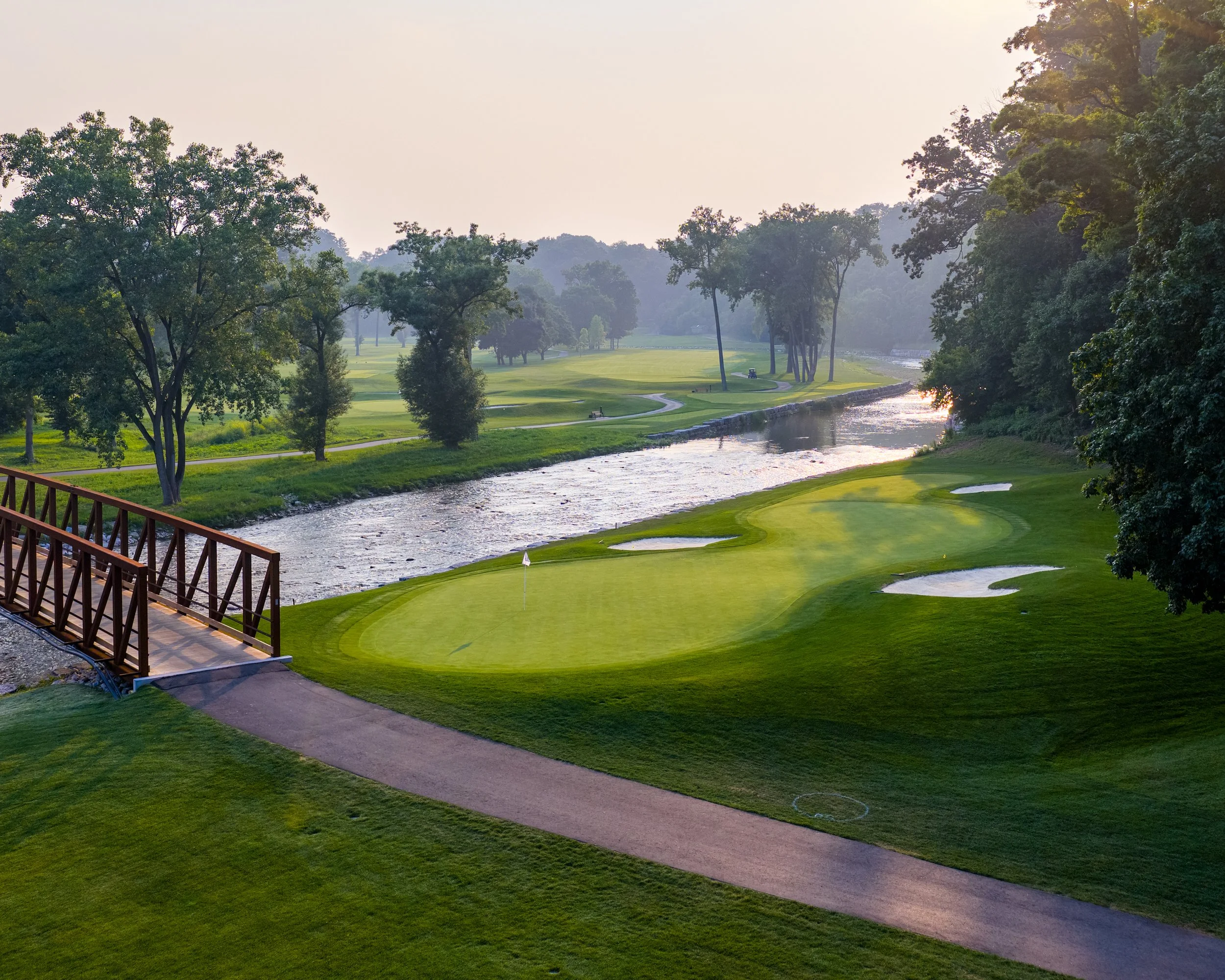 A scenic golf course with green fairways, water hazard, and a row of trees. A wooden railing is visible in the foreground, and the sky is overcast.