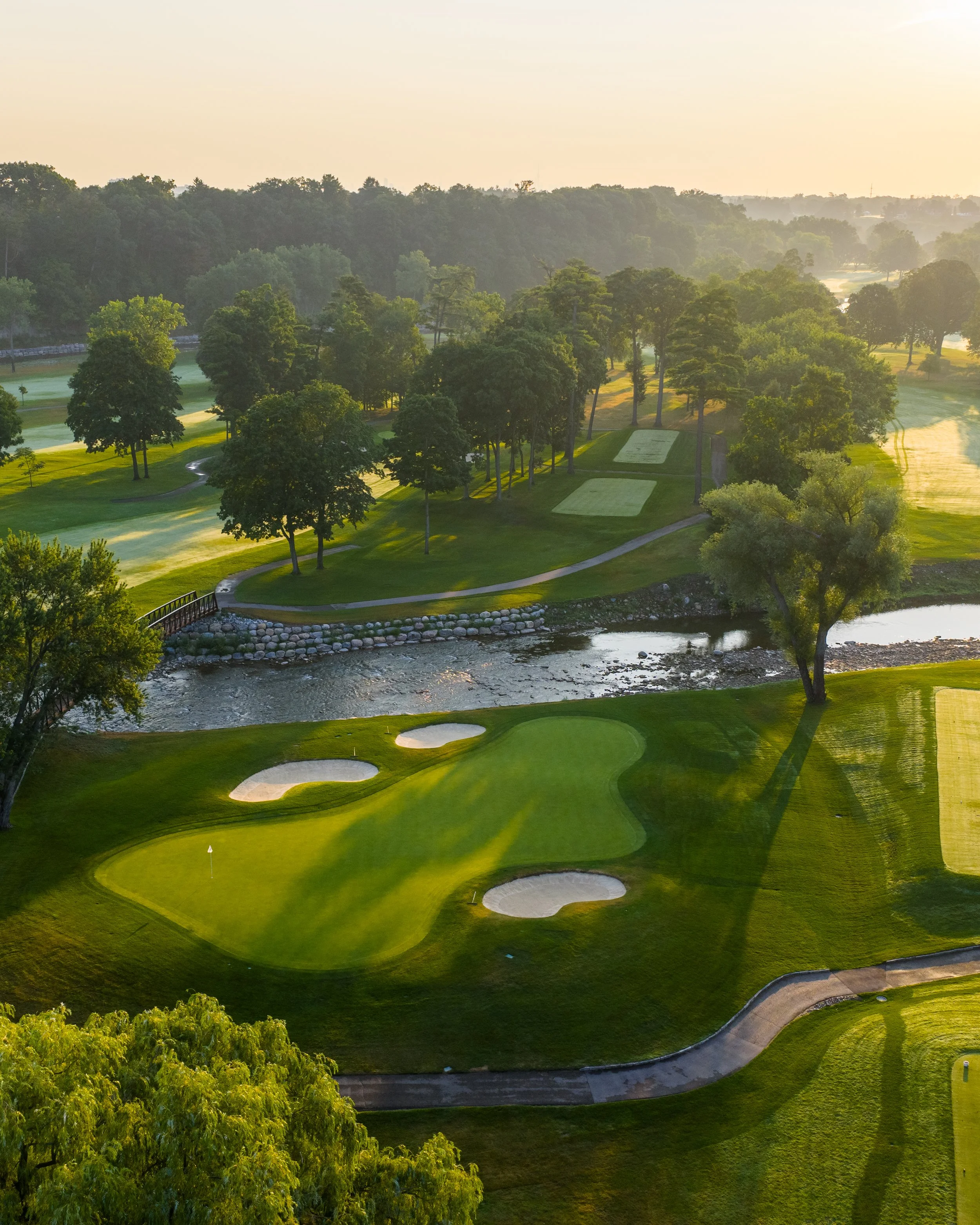 Aerial view of a golf course with lush green grass, sand bunkers, water hazards, and a surrounding forest under a hazy sky at sunrise or sunset.
