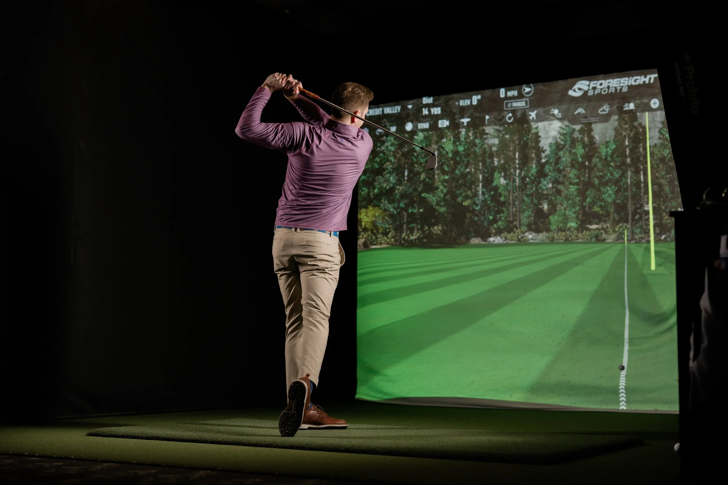 A man in a purple long-sleeve shirt and khaki pants is swinging a golf club inside a simulation room with a large screen showing a golf course.