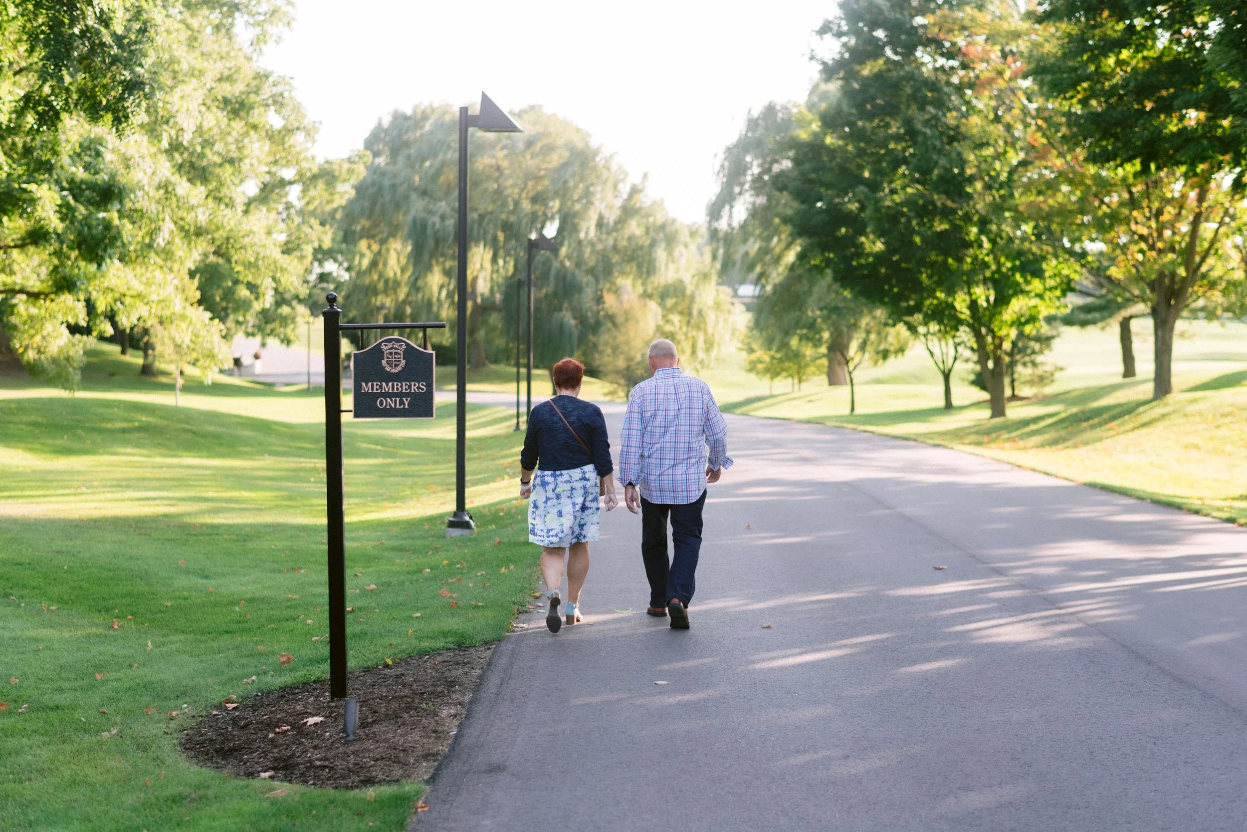 An elderly couple walking together in a park on a sunny day, with a sign reading "Members Only" nearby. The park is lush with green trees and grass.