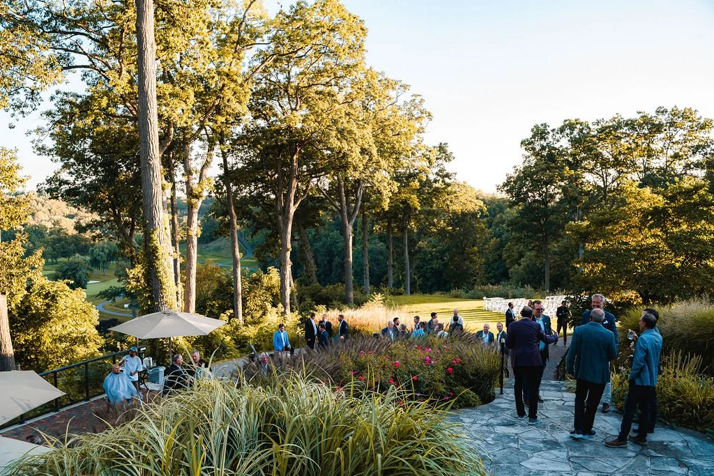 People gathering outdoors on a stone-paved patio surrounded by lush greenery and tall trees during late afternoon or early evening.
