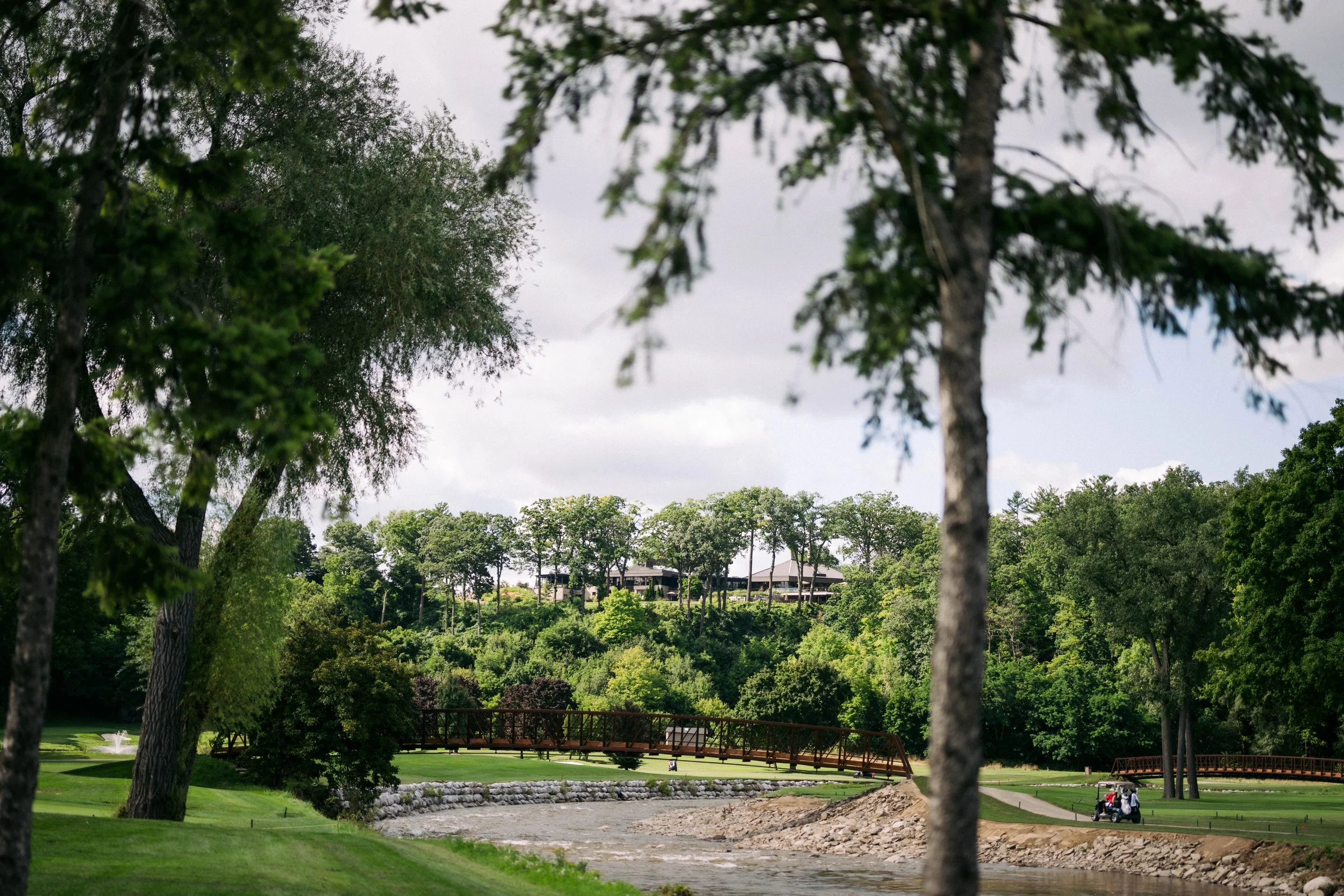 A lush green golf course with a small bridge over a pond, surrounded by trees, and houses in the distance under a cloudy sky.
