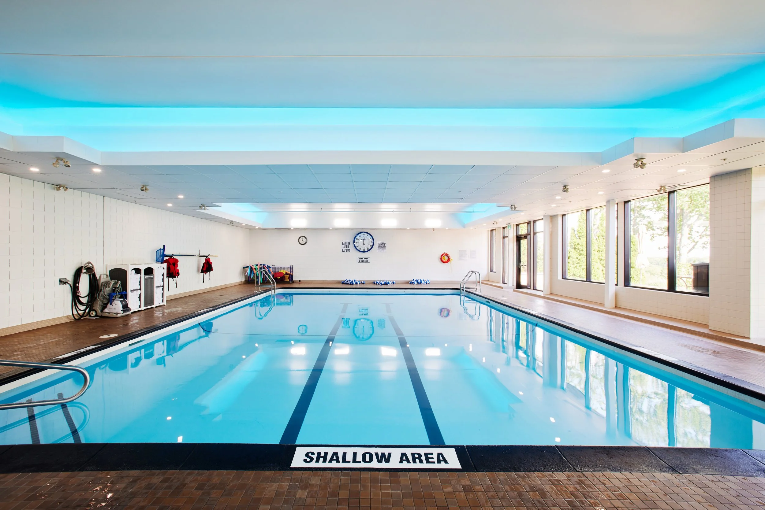 Indoor swimming pool with a sign that reads 'Shallow Area' at the edge. The pool has clear water, poolside vacuum equipment, life preserver, and colorful pool noodles. The room has large windows with trees outside, a white tiled wall, and a ceiling with blue lighting accents.
