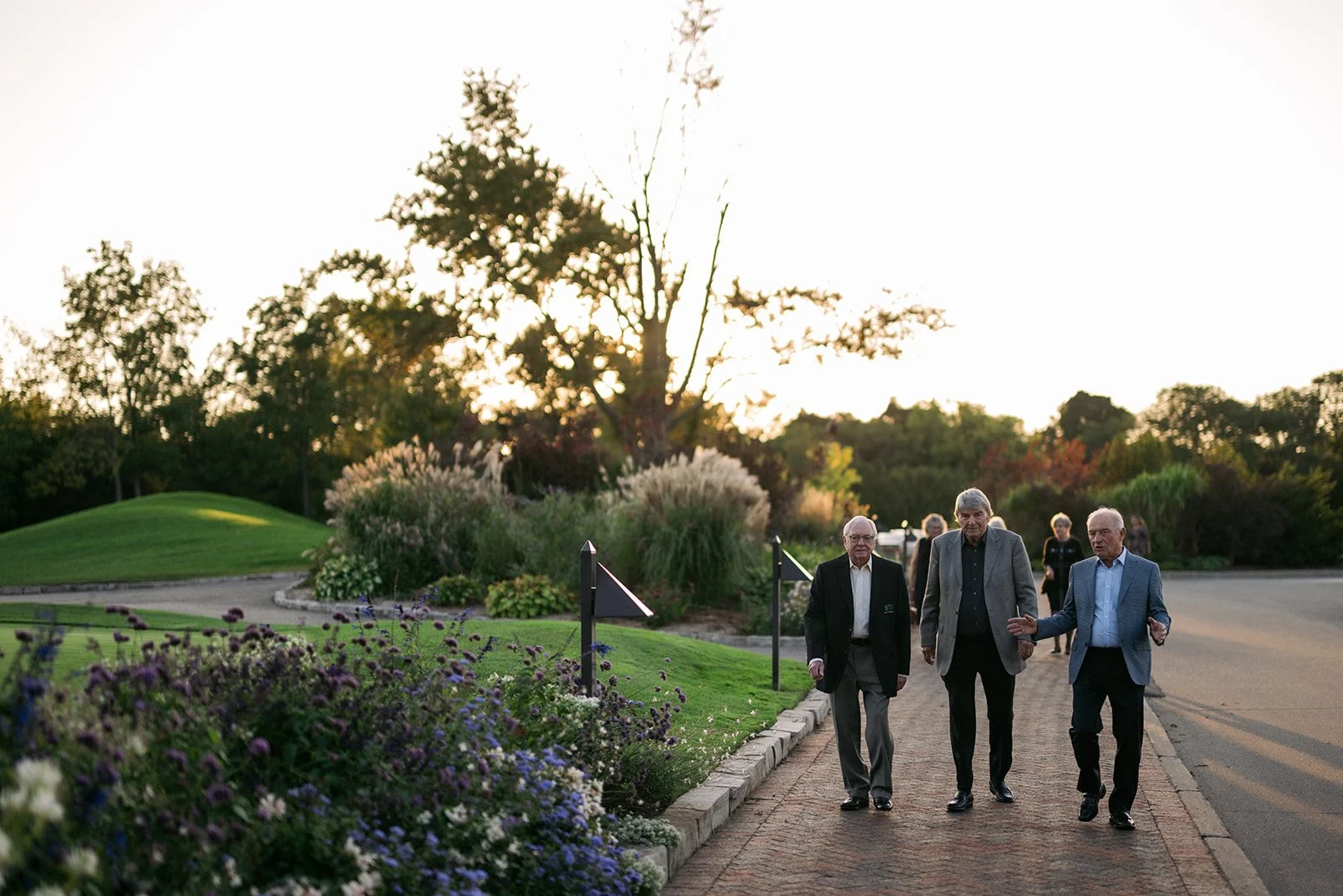 Three elderly men walking together on a brick sidewalk in a park-like area with greenery, trees, and flowers during sunset.