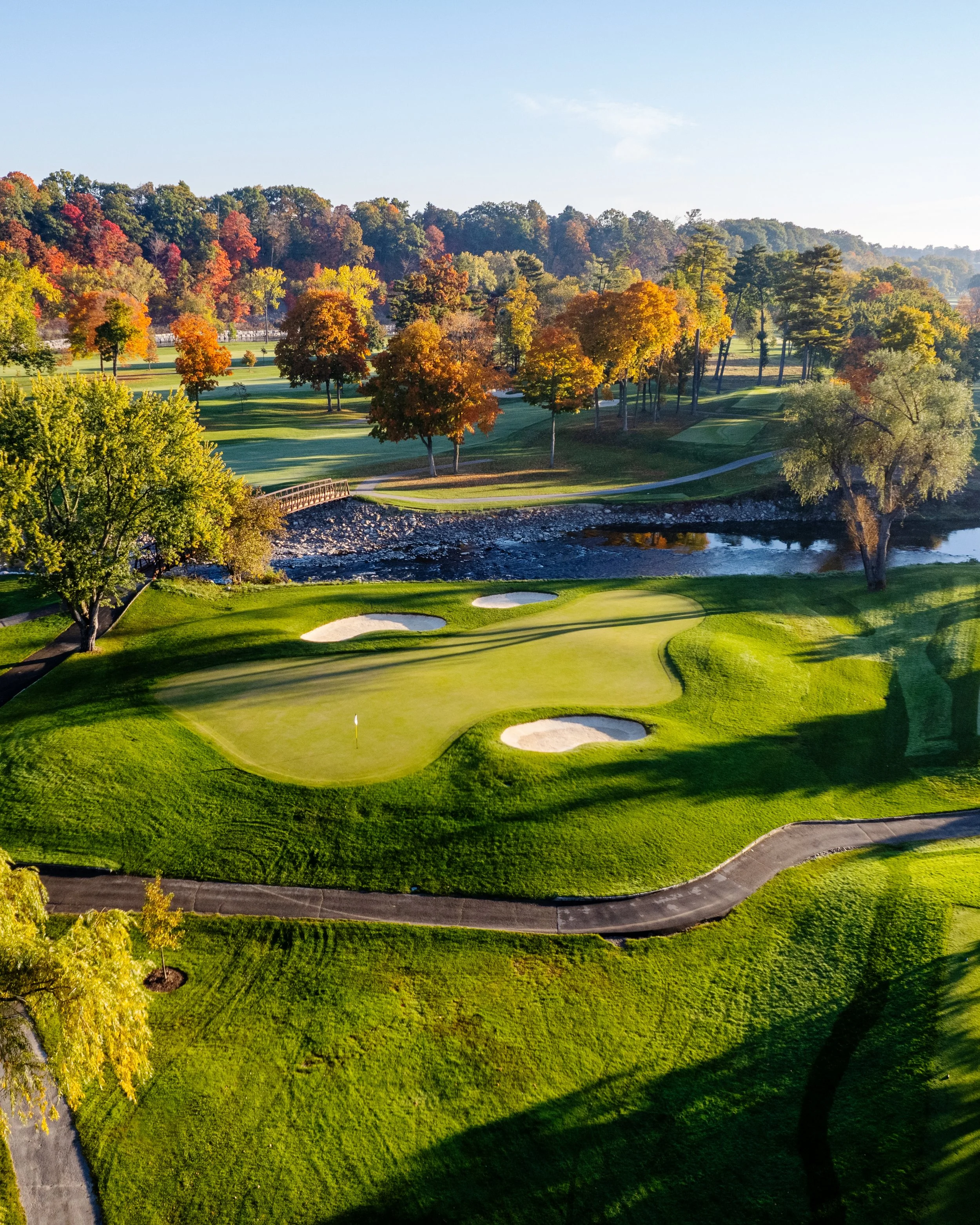 A scenic view of a golf course surrounded by colorful trees during autumn, with a small river flowing along the course and clear skies overhead.