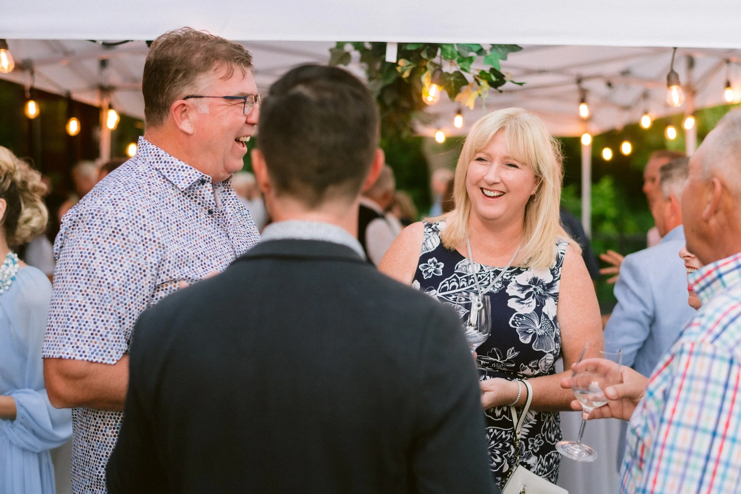People at an outdoor party or social gathering, smiling and conversing, under string lights and a canopy, with greenery in the background.