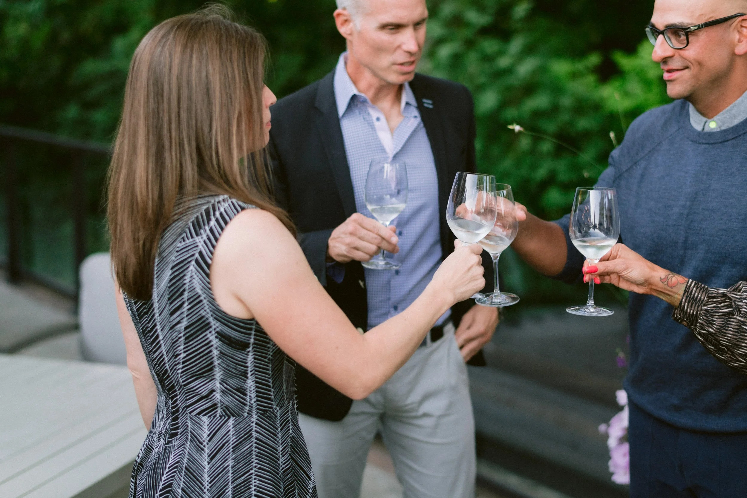 Group of four people holding wine glasses at an outdoor gathering with greenery in the background.