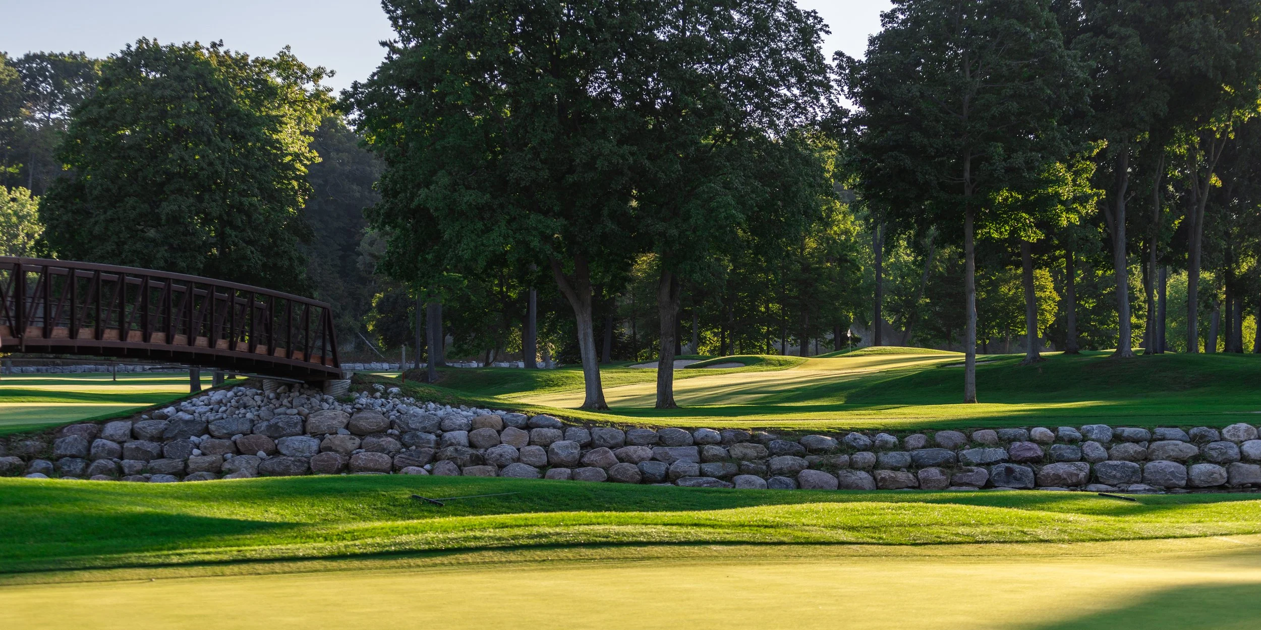 A view of a golf course with a small bridge over rocks, lush green trees, and well-maintained grass.