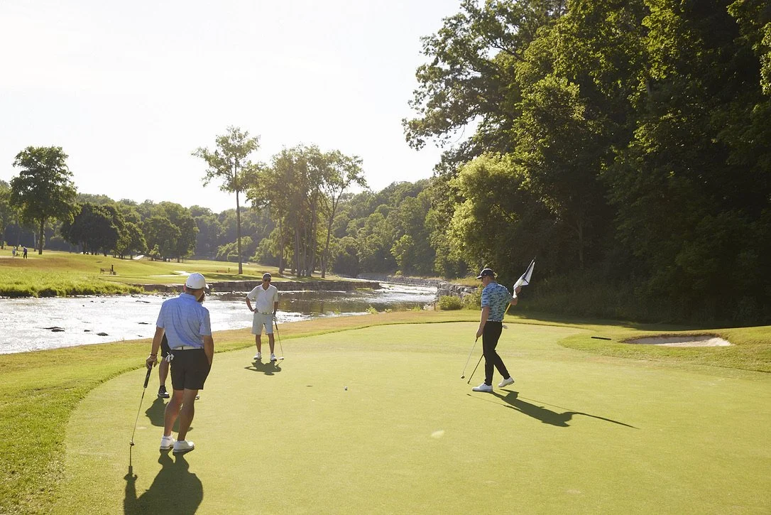 Four men play golf on a green fairway near a river, with trees in the background on a sunny day.