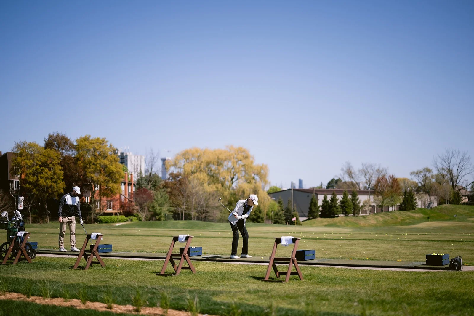 Two people practicing golf at a driving range on a sunny day, with trees and buildings in the background.