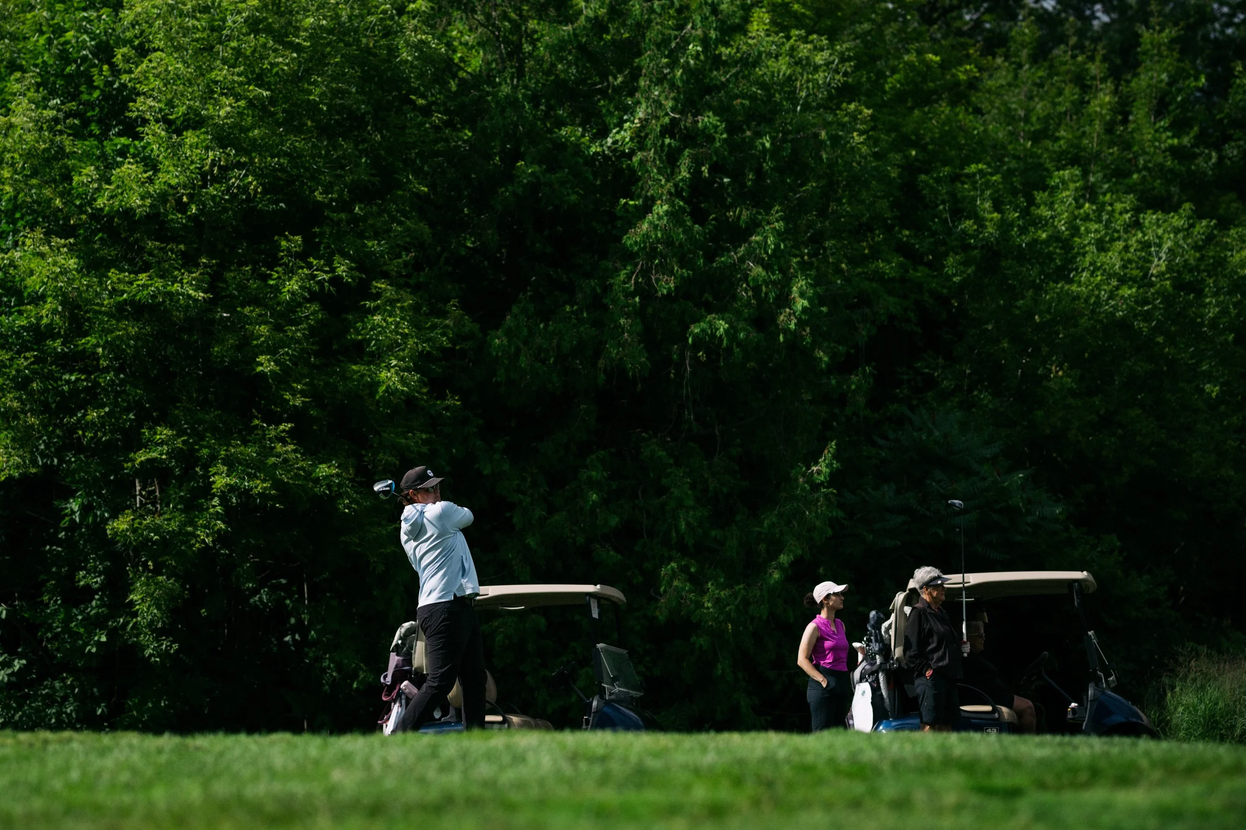 Group of people at a golf course, some standing beside golf carts, with lush green trees in the background.