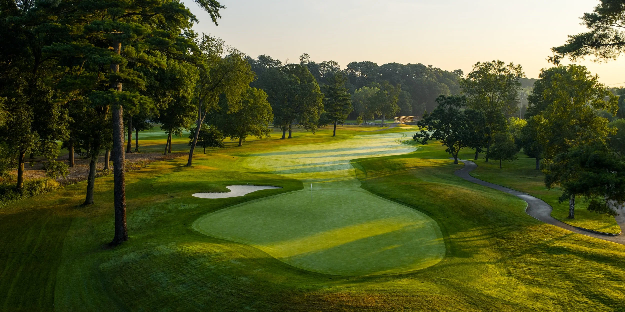A lush golf course with green fairways, trees lining the course, and a small pond near the hole with a flagstick. The scene is illuminated by warm sunlight, creating long shadows.