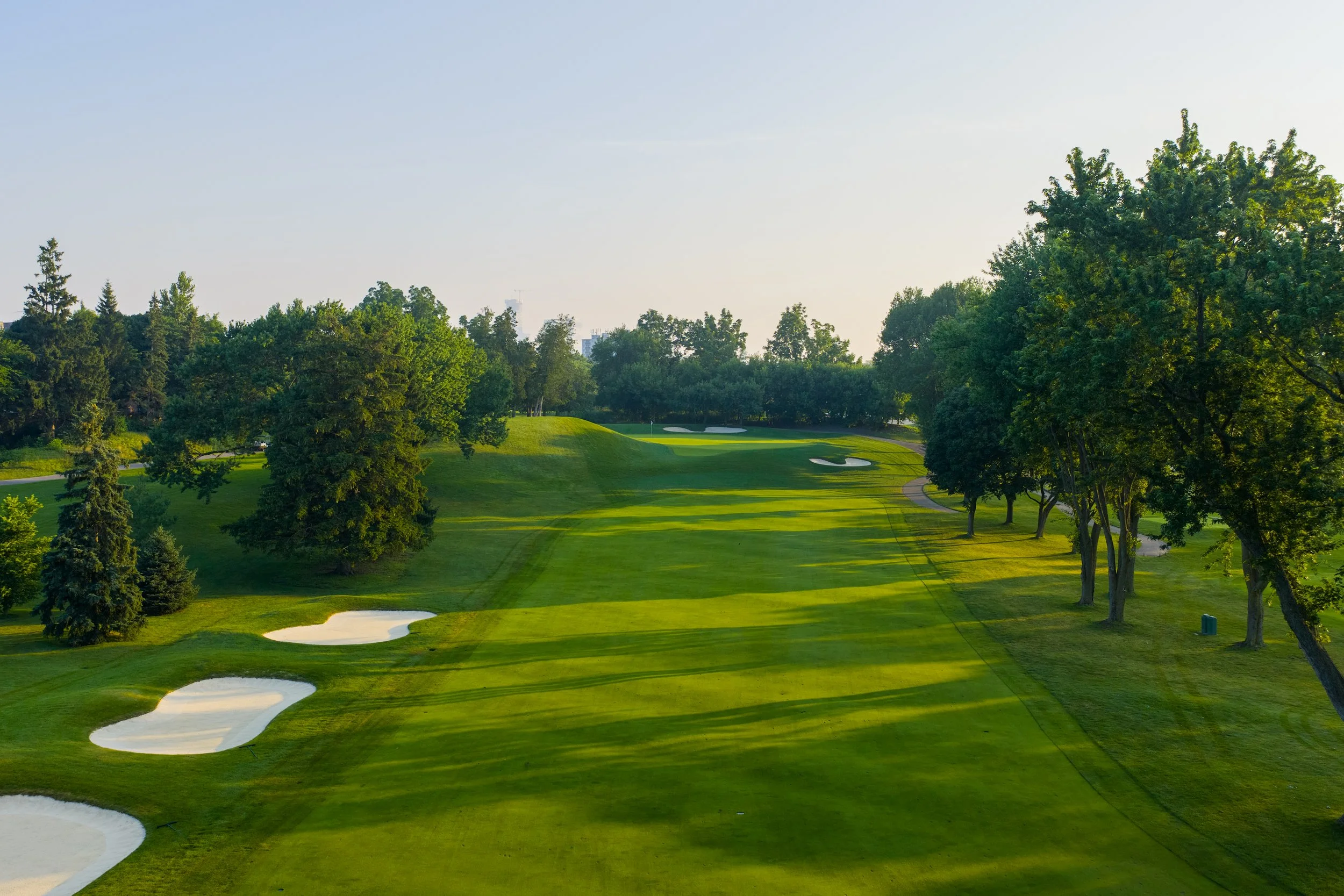 A green golf course with sand bunkers and tall trees, under a clear sky.