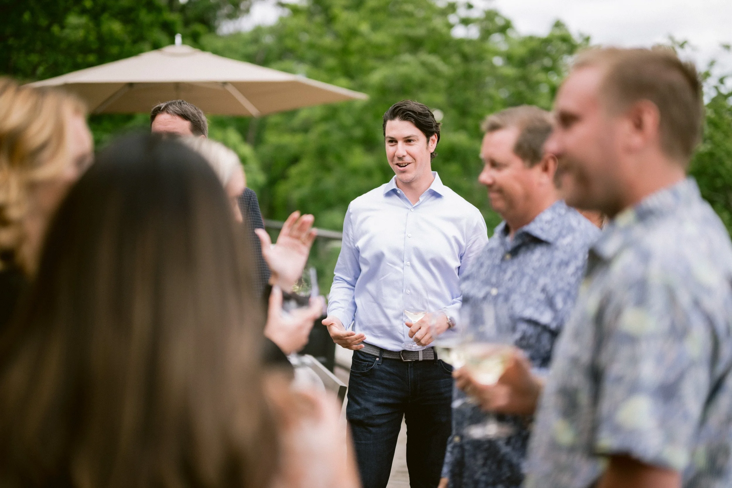 Group of people at an outdoor event, with one man in a light blue shirt speaking or socializing amidst others, under a large umbrella with green trees in the background.