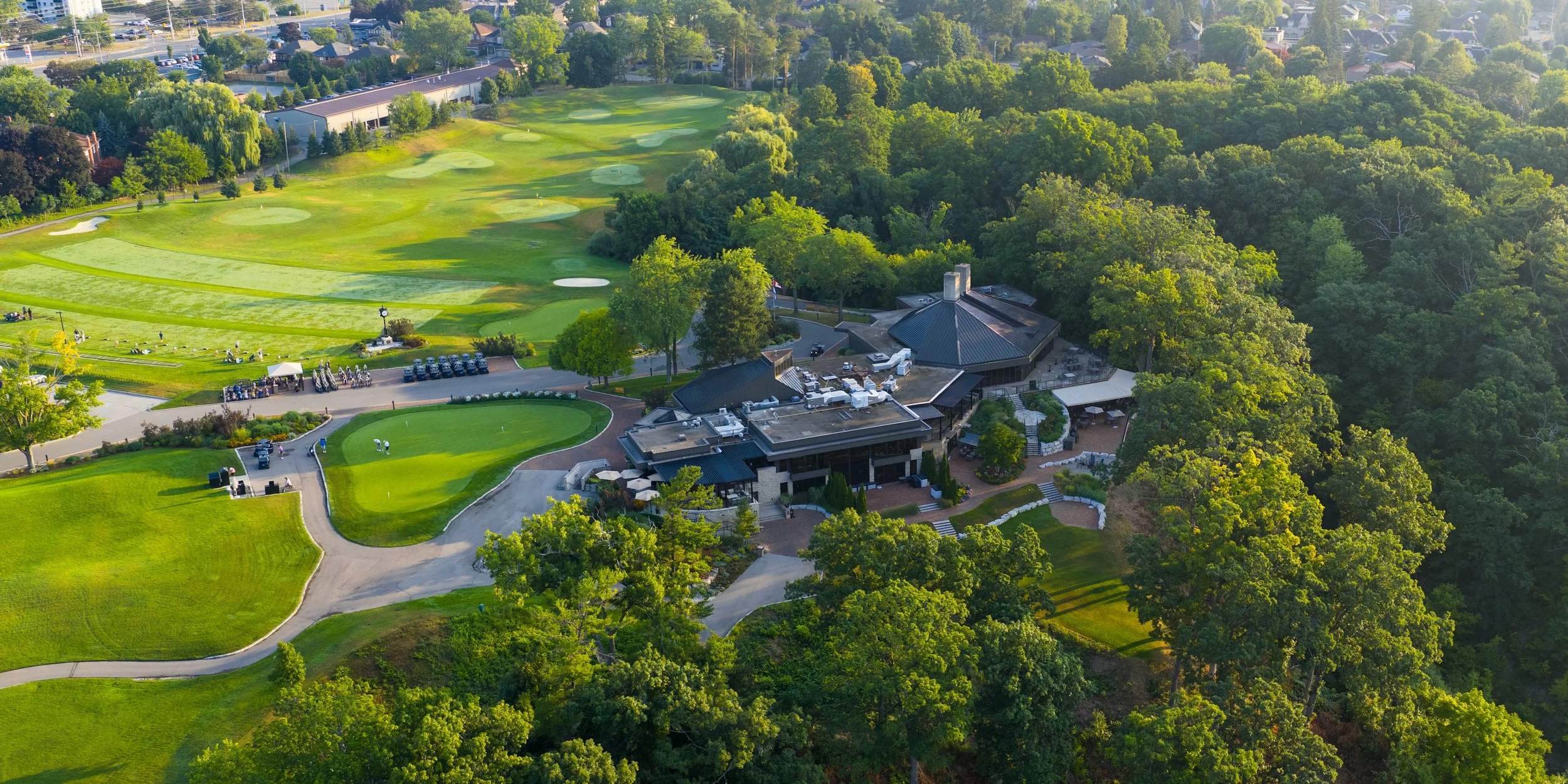 Aerial view of a golf course with lush green fairways and trees, a clubhouse with outdoor seating, parking lot, and surrounding residential neighborhoods.