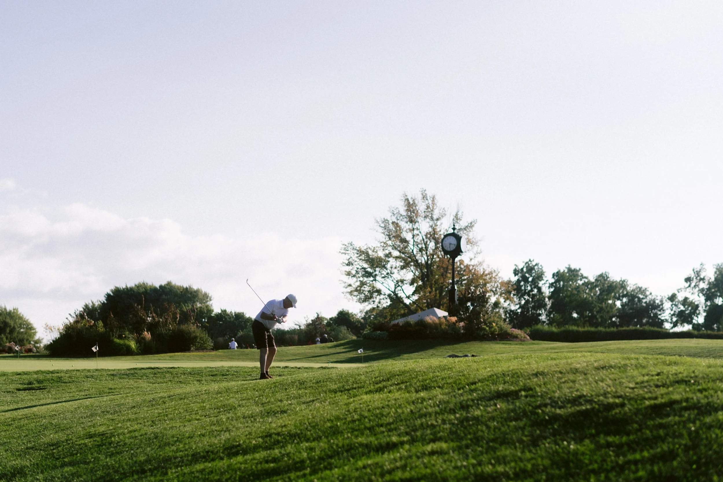 A golfer preparing to putt on a golf course with lush green grass, trees, and a clock on a pole in the background under a partly cloudy sky.