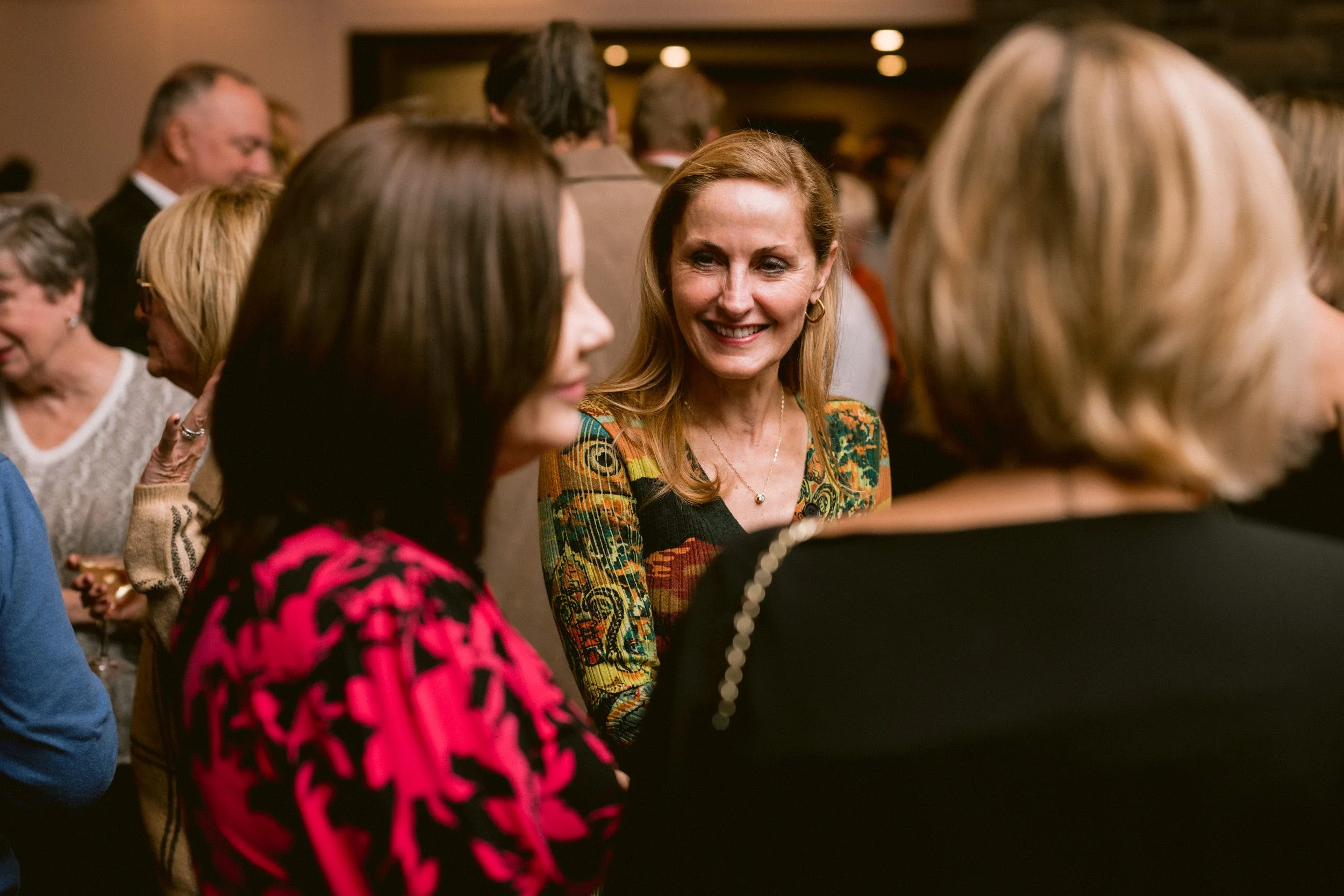 Group of people socializing at an indoor event, with the woman in the center smiling at others.