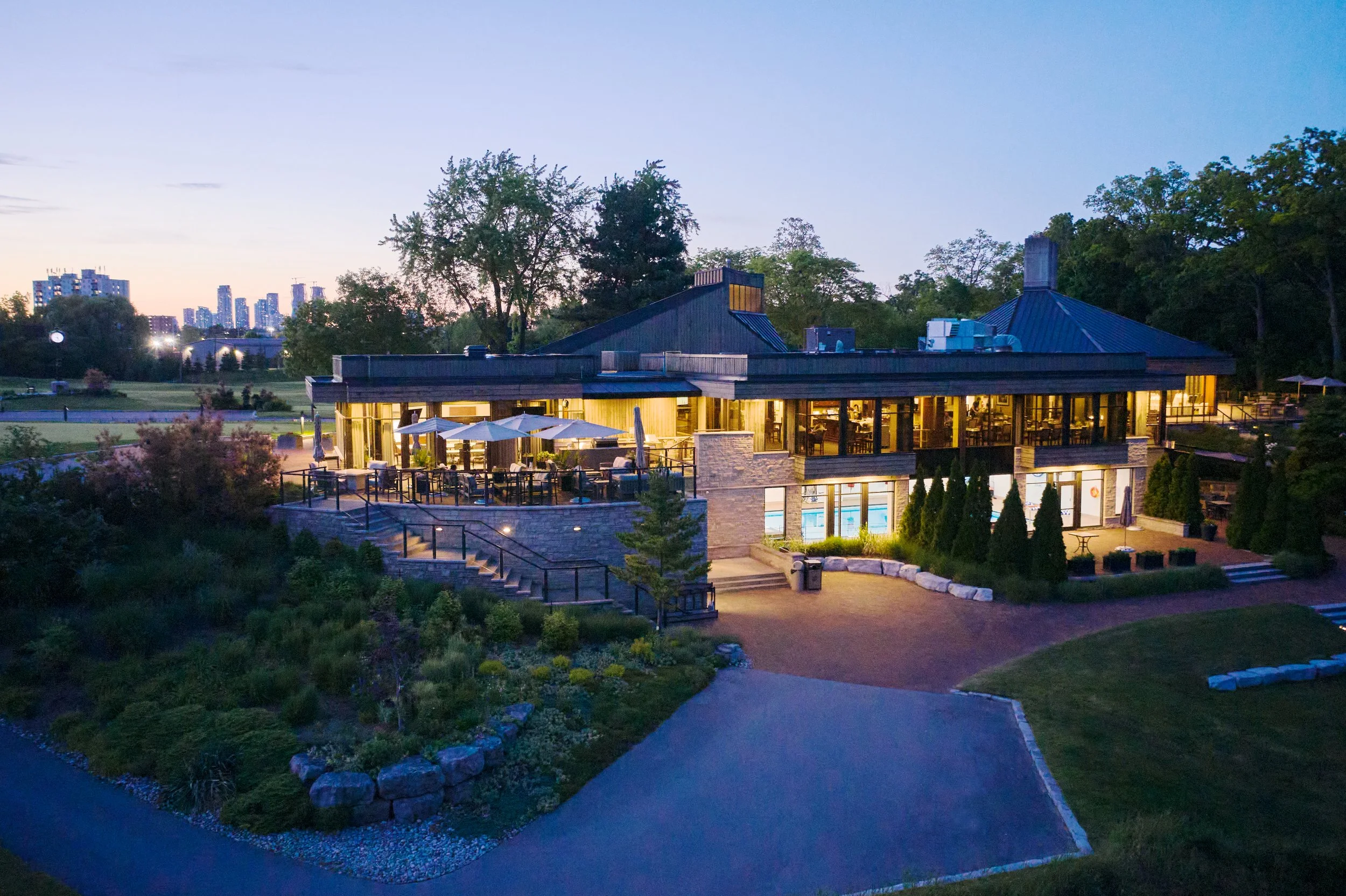 A peaceful evening view of a modern building with large glass windows, surrounded by greenery and trees. Warm lights glow from inside, and a patio with umbrellas and tables is visible. In the background, a city skyline with tall buildings is seen aga
