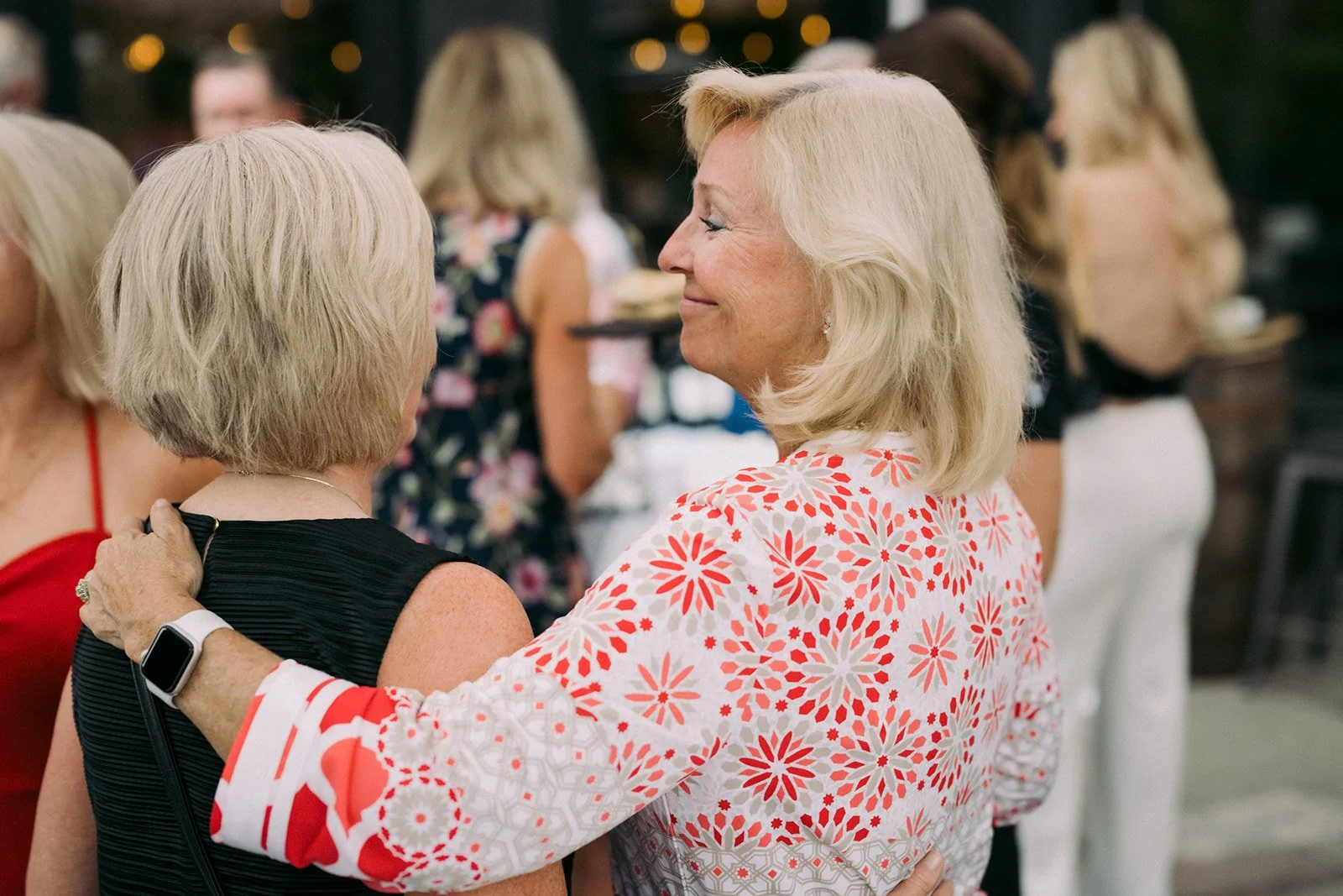 Two older women are in a social setting, smiling and holding each other. The woman on the right has blonde hair and is wearing a white shirt with a red and gray floral pattern. The woman on the left has short blonde hair and is wearing a black sleeve