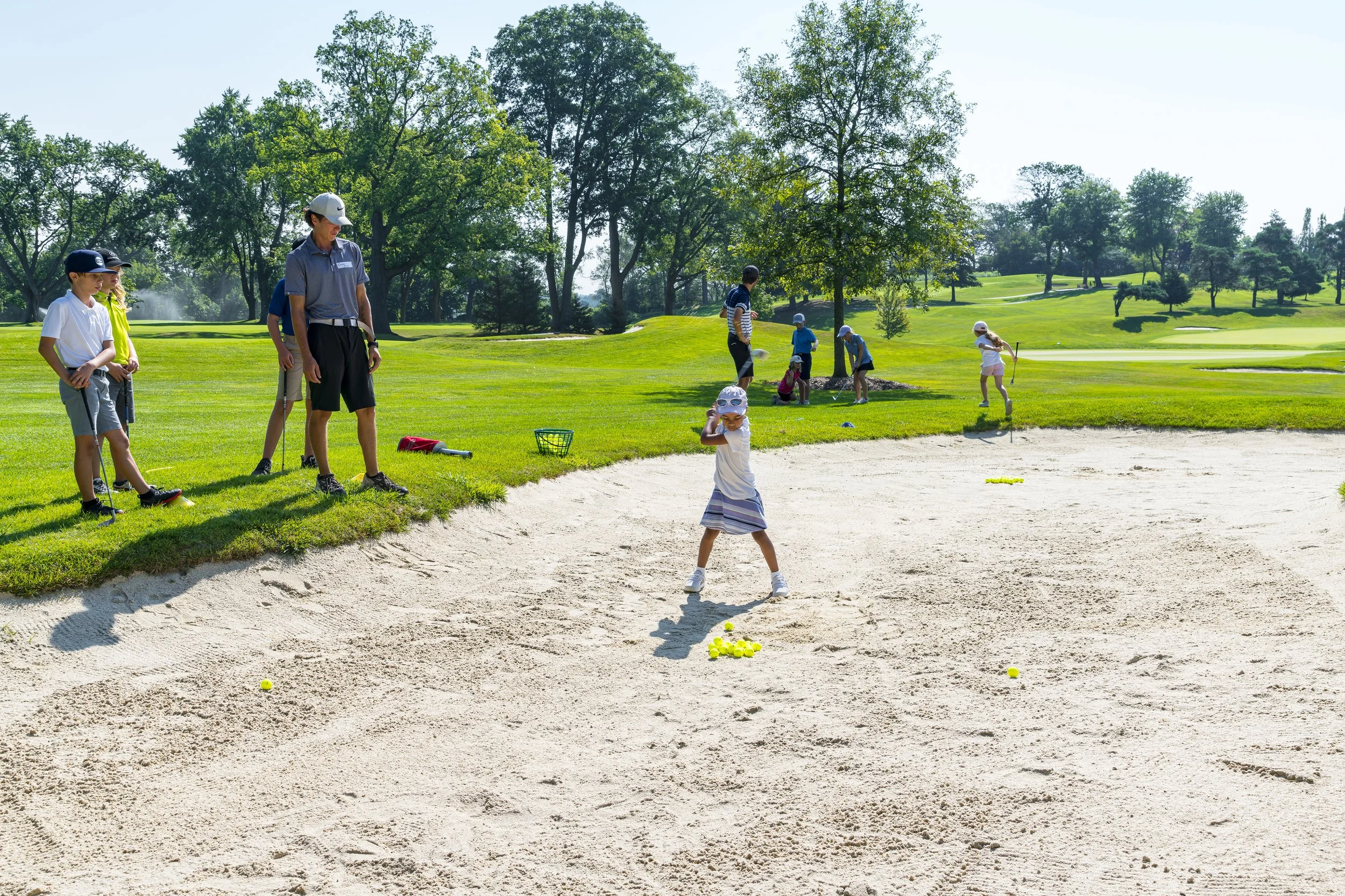 Children and adults playing and practicing golf in a sand trap at a golf course on a sunny day.