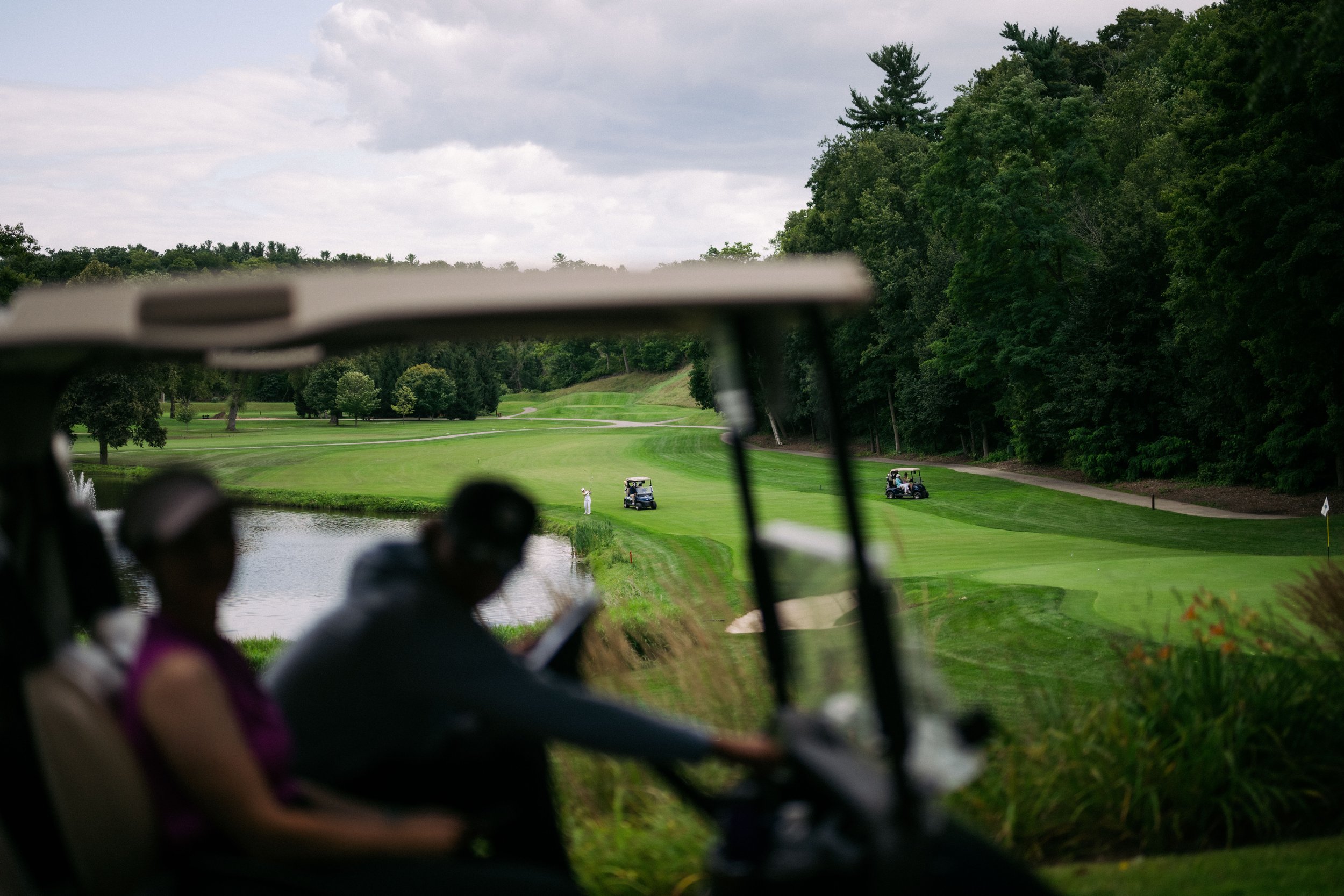 Golf course with golfers, golf carts, water hazards, and lush trees under a cloudy sky, viewed from a golf cart.