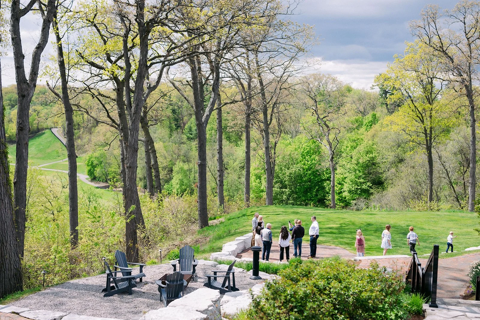 People gathered on a grassy area in a park with tall trees and a winding path in the background, under a cloudy sky.