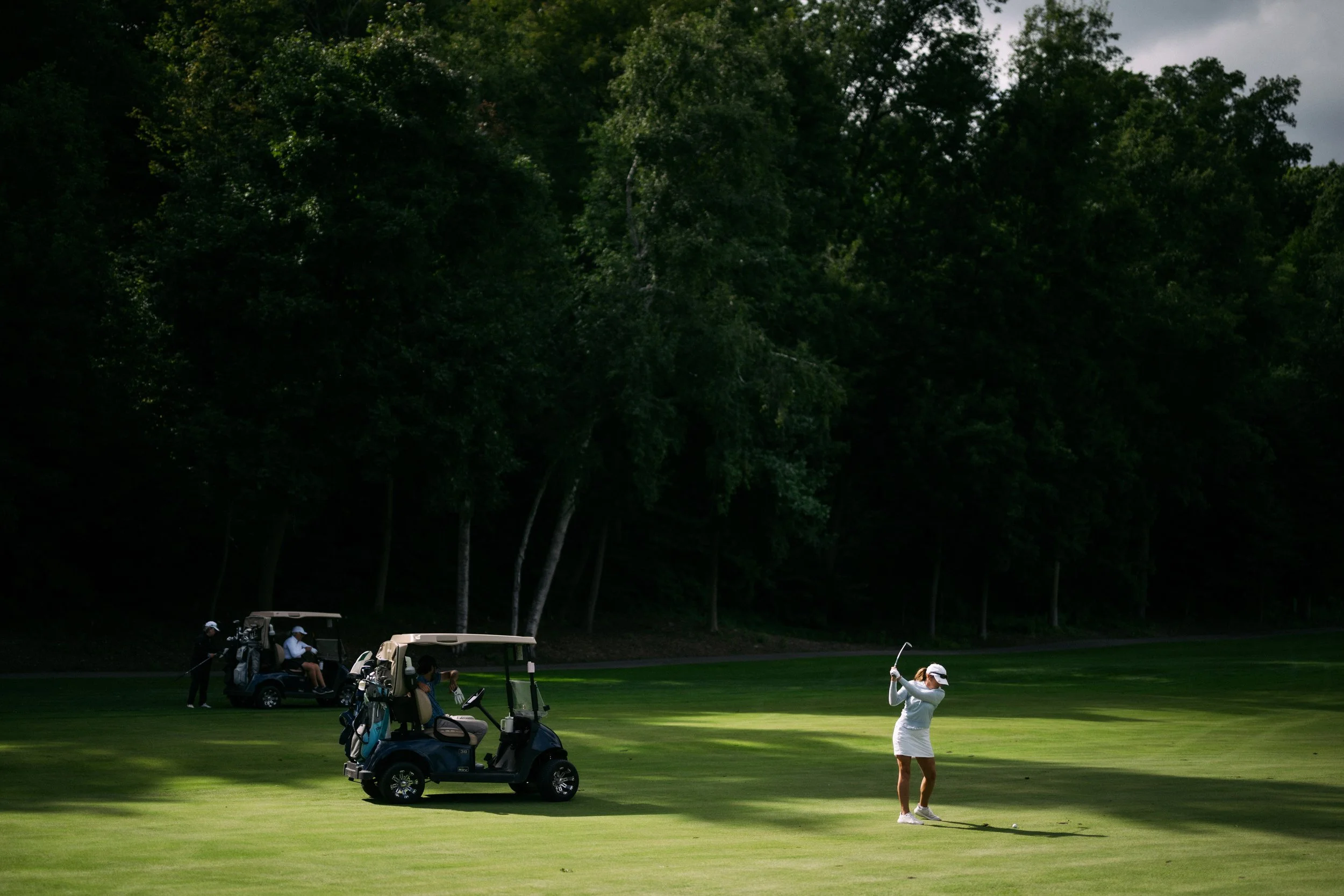 A woman playing golf on a lush green golf course, with several golf carts and people in the background, surrounded by trees.