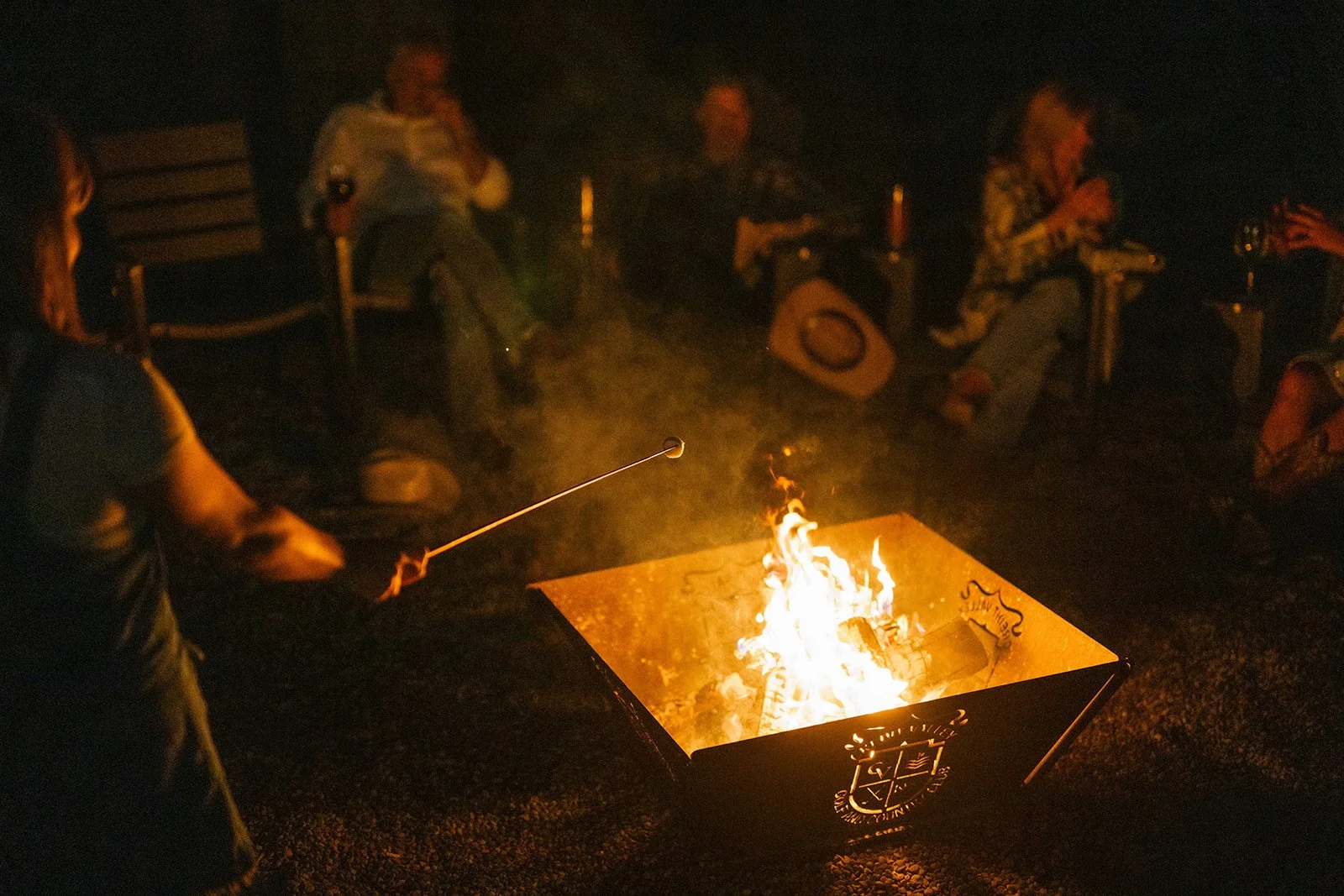 People sitting around a fire at night, one person is roasting marshmallows on a stick, and others are relaxing.