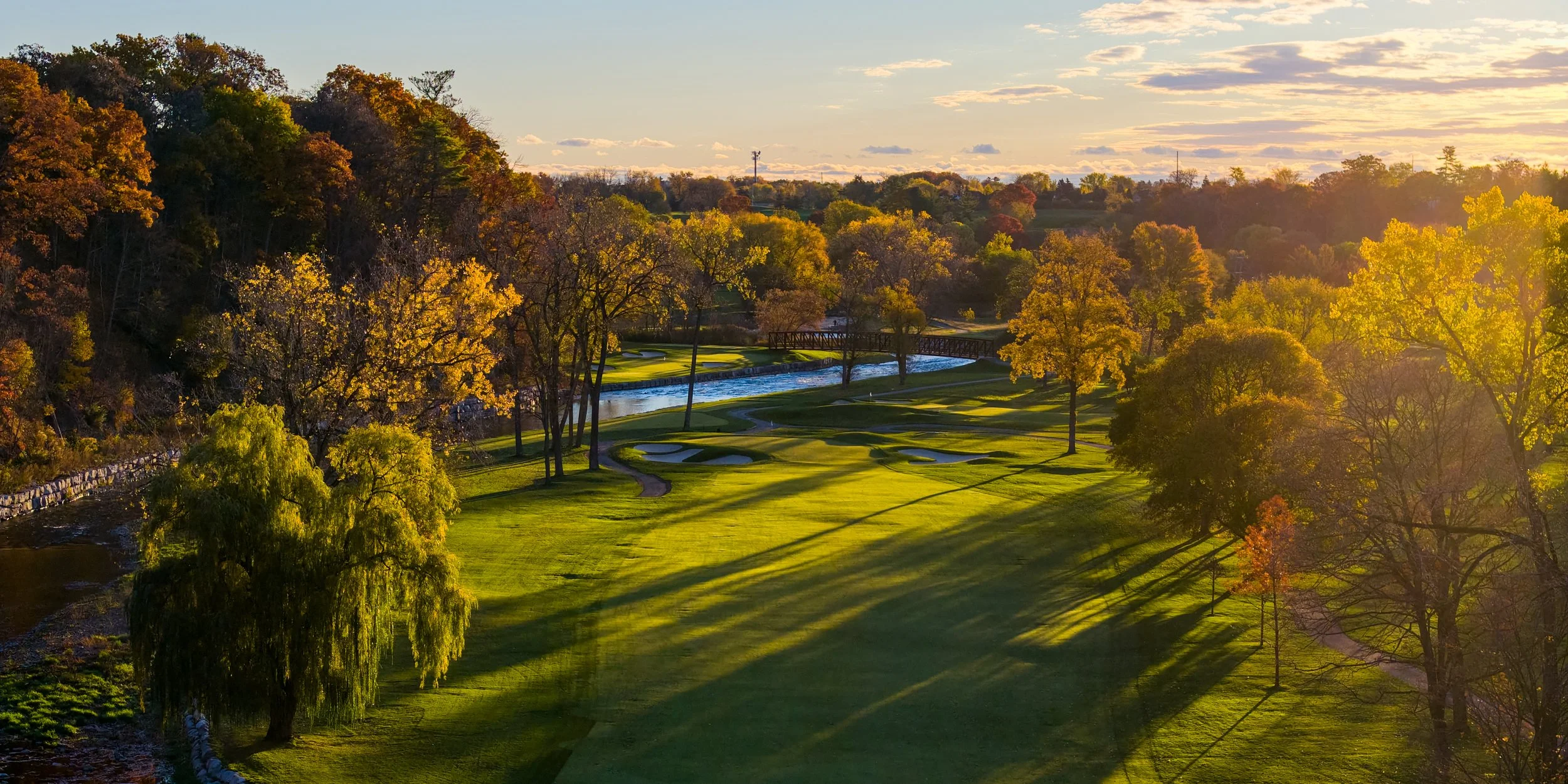 A scenic view of a golf course surrounded by trees with autumn leaves, a river, and a bridge, under a partly cloudy sky with the sun casting shadows.