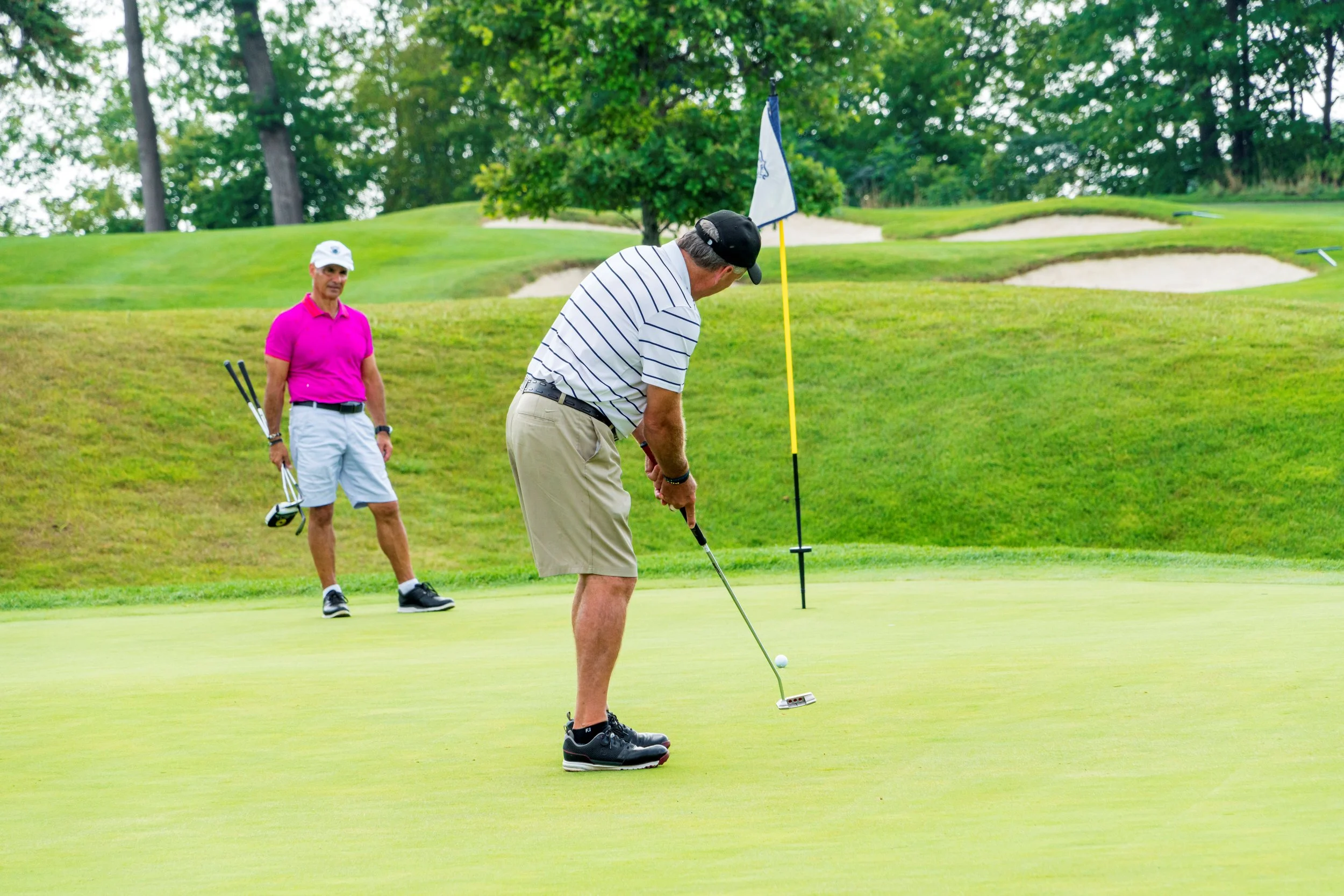 Two men playing golf on a lush green golf course. One man is preparing to putt, while the other holds golf clubs and observes. There are trees and sand bunkers in the background.