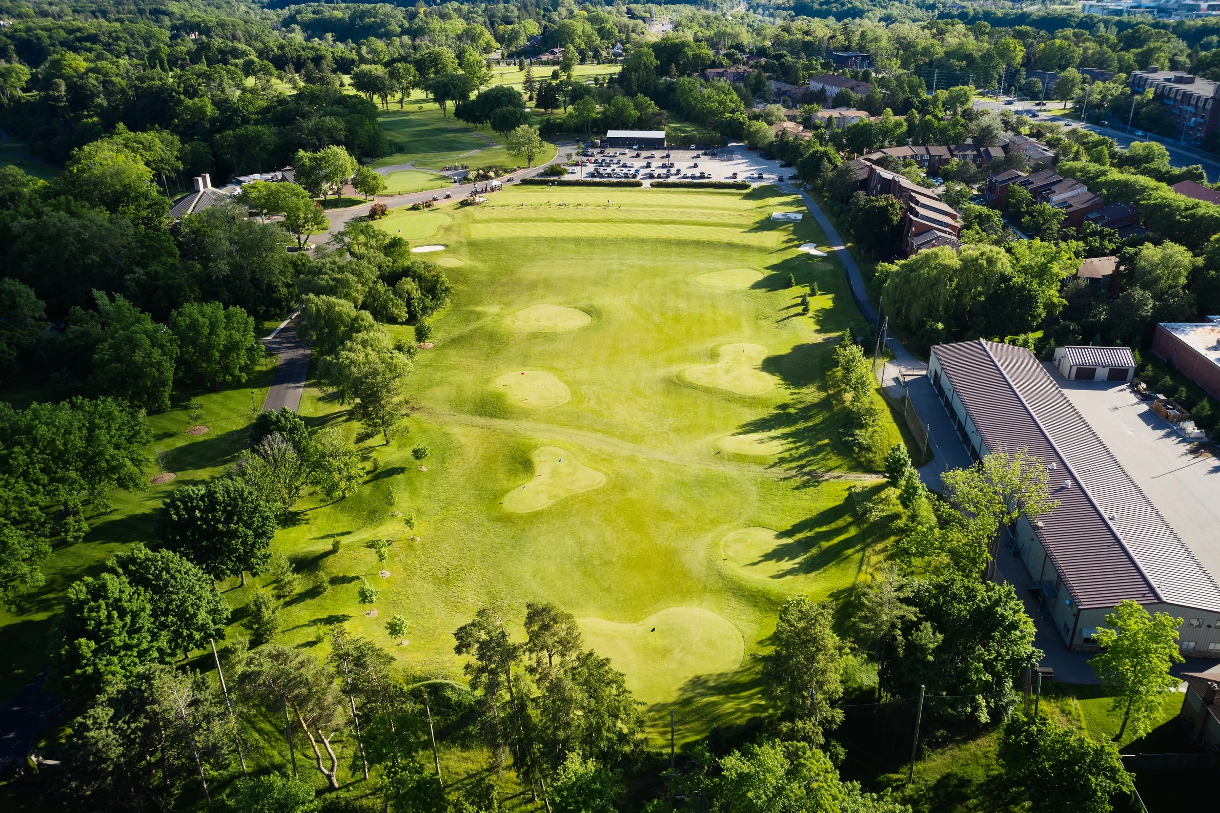 Aerial view of a golf course with green fairways, sand traps, surrounded by trees, adjacent to parking lot and buildings.