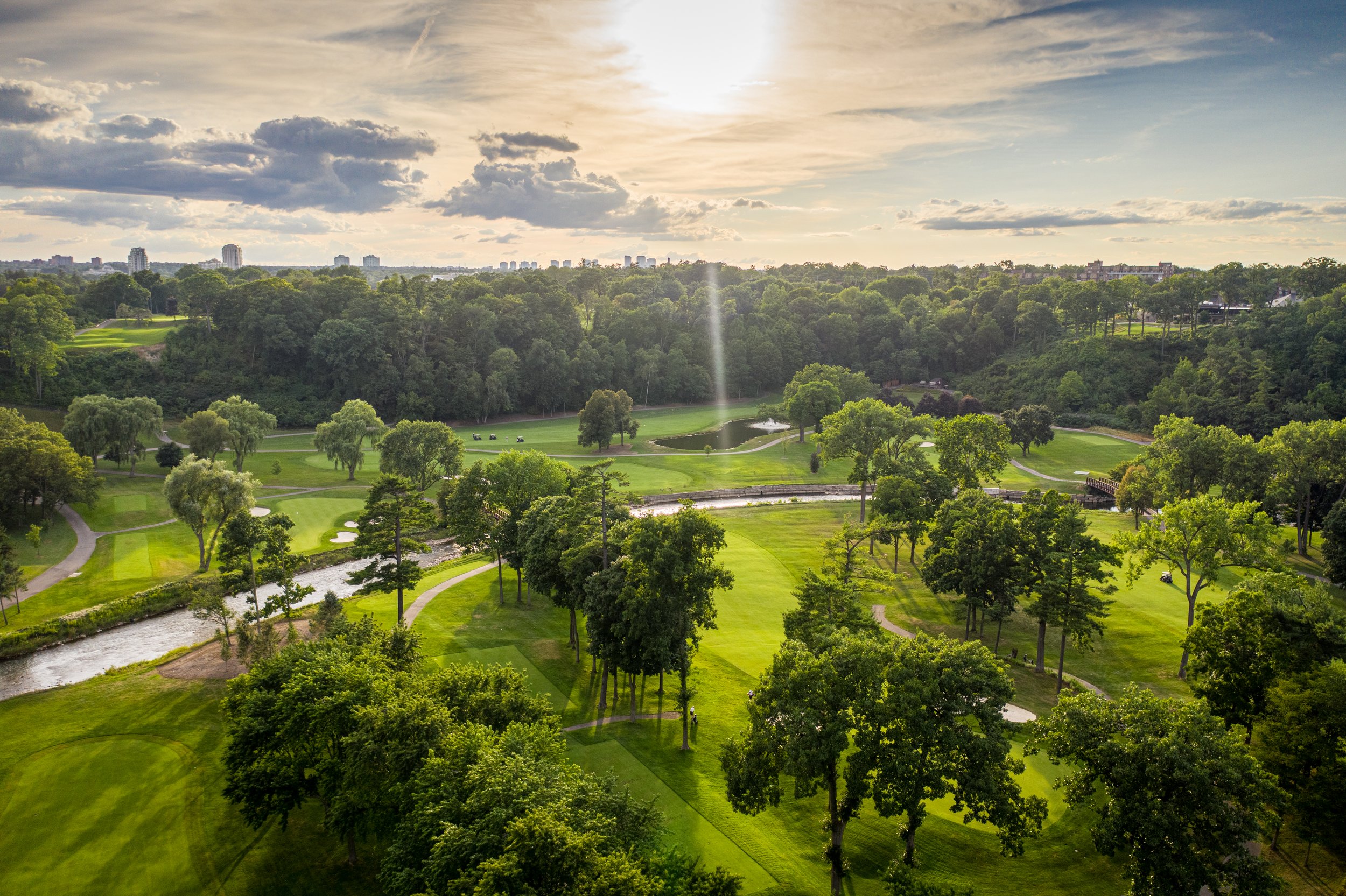 A scenic view of a golf course surrounded by trees, with a pond, walking paths, and a city skyline in the distance under a partly cloudy sky at sunset.