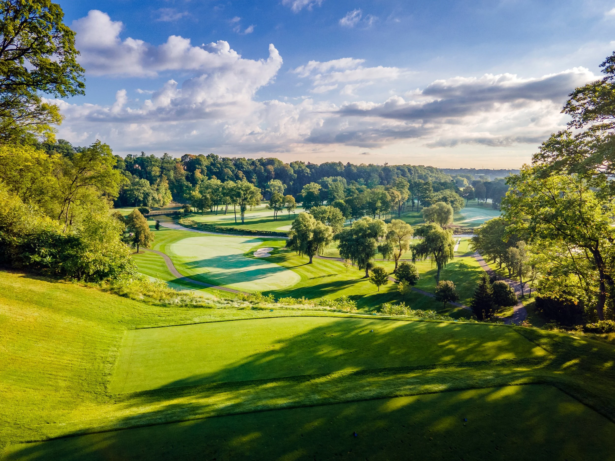 A scenic view of a golf course surrounded by lush green trees, with a partly cloudy sky overhead.
