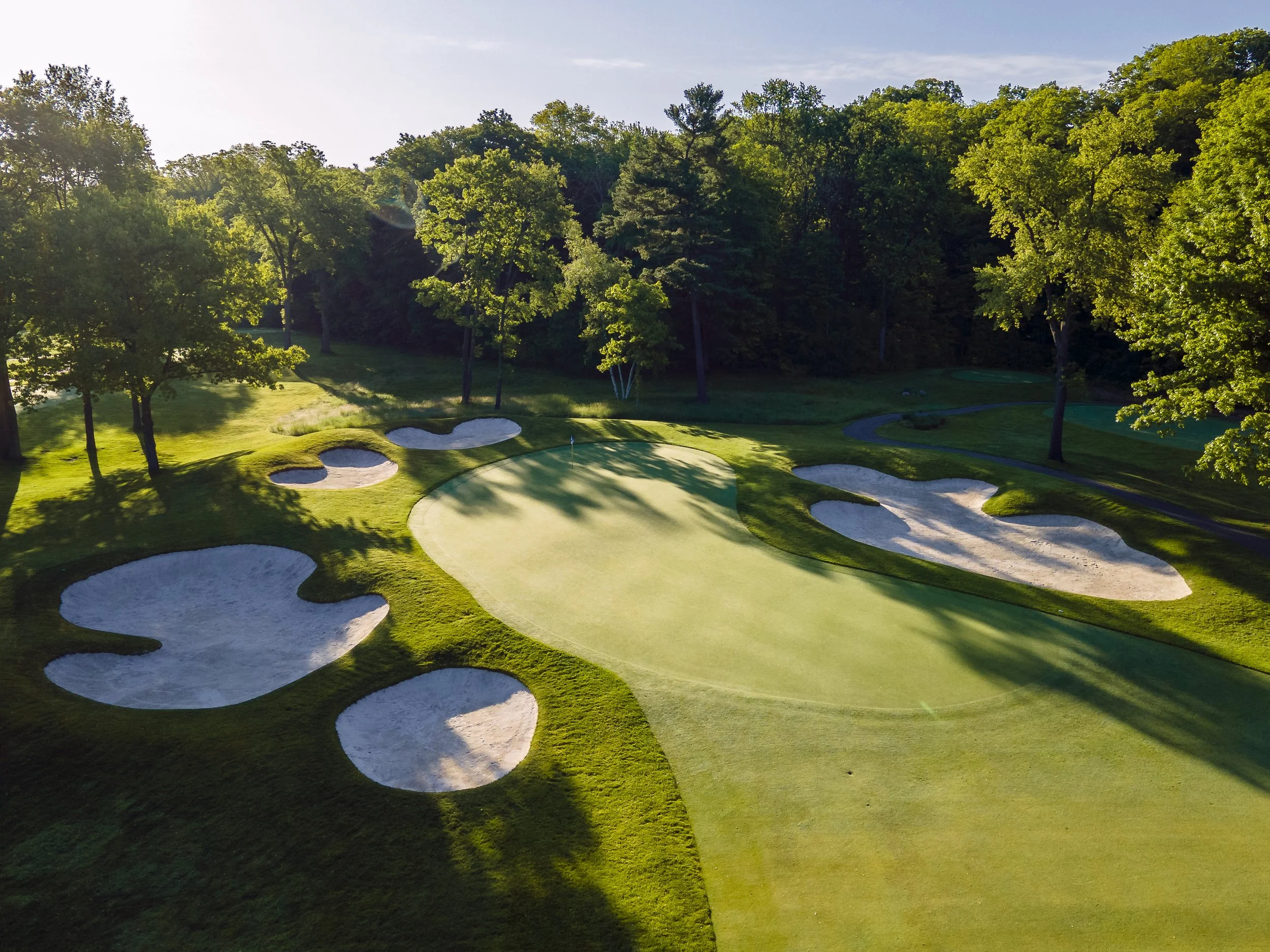 A golf course with neatly maintained greens, sand bunkers, and surrounding tall trees on a sunny day.