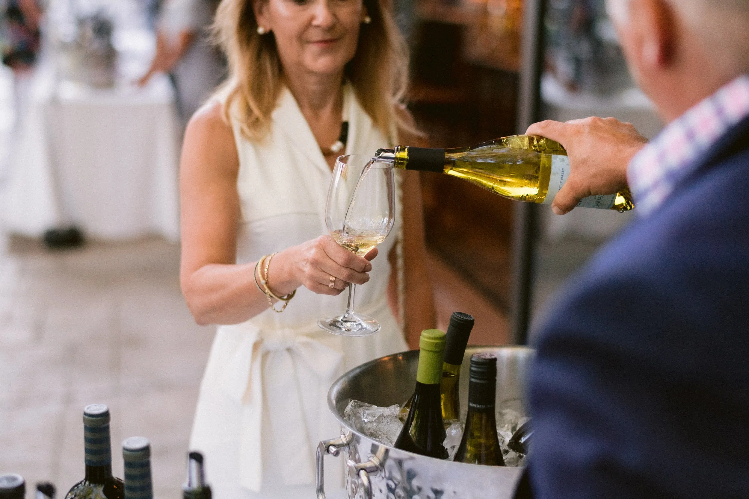 A woman in a white dress is served white wine into a glass at a wine tasting event, with bottles of wine on ice in a bucket in front of her.