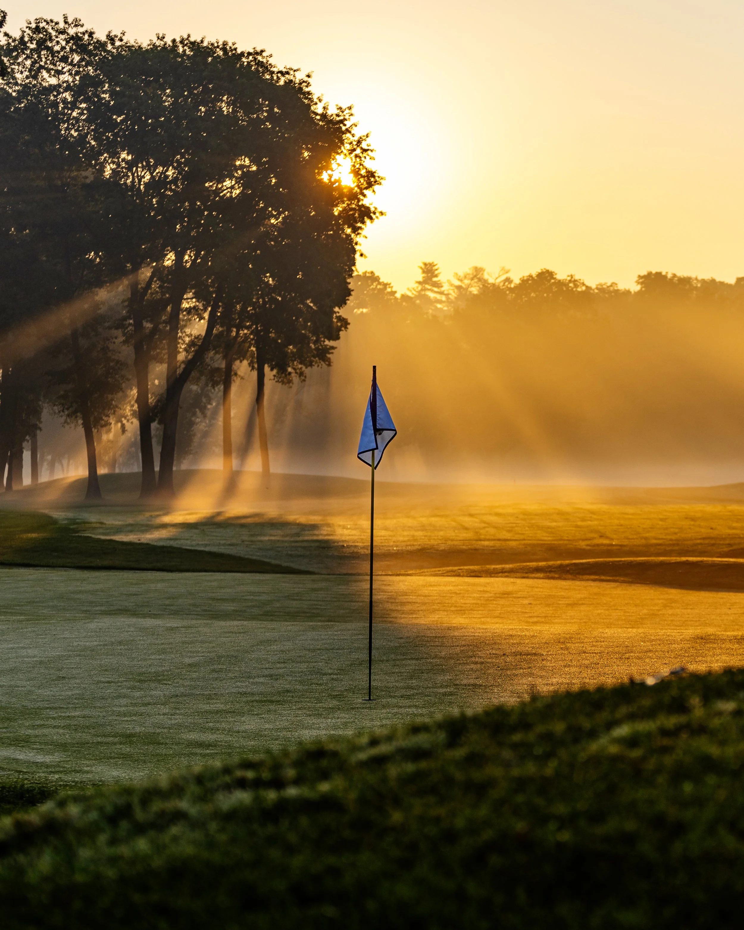 Sunrise over a golf course with trees and a flag on the green.