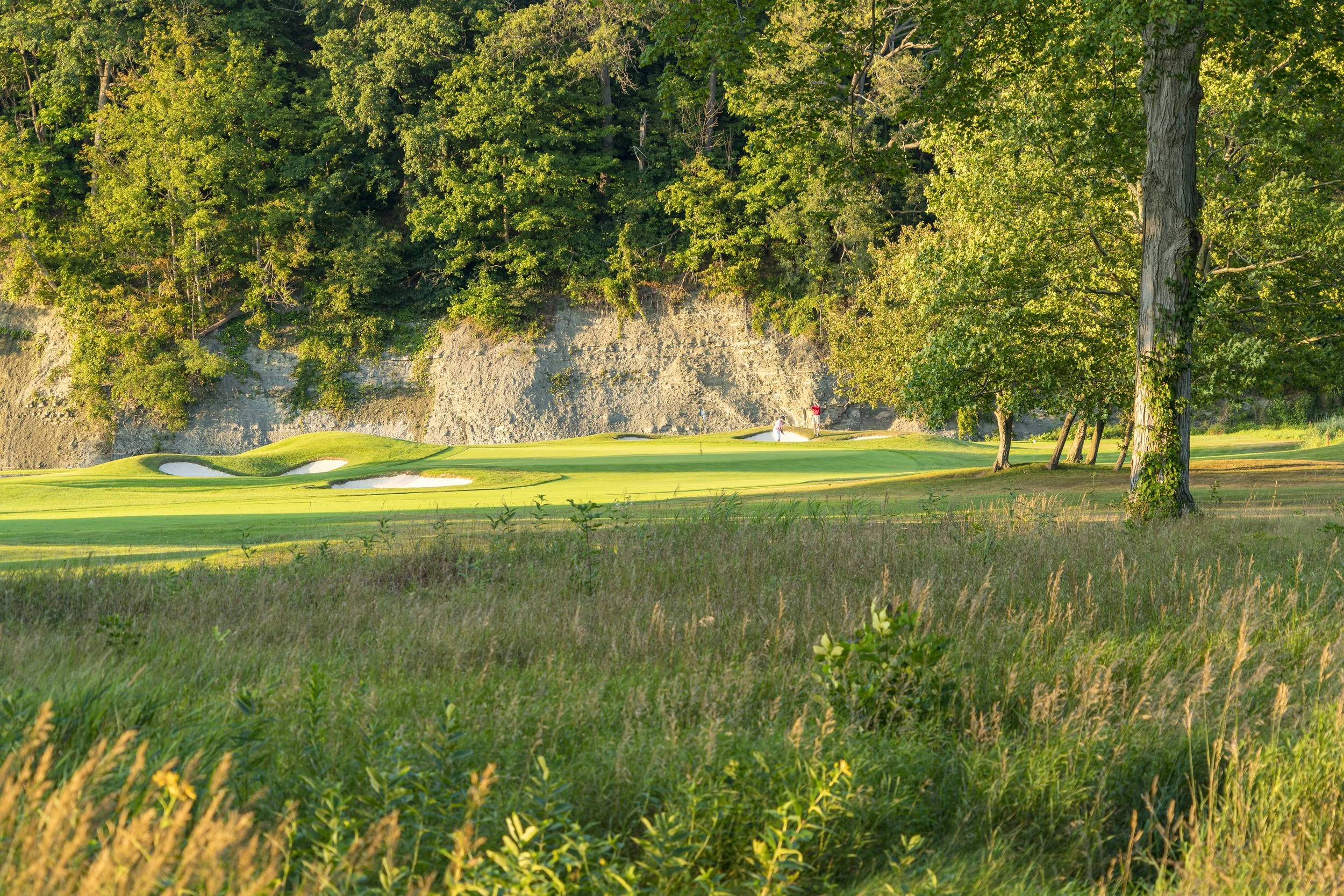 A golf course with two people playing near a wooded area and a rocky hill in the background.
