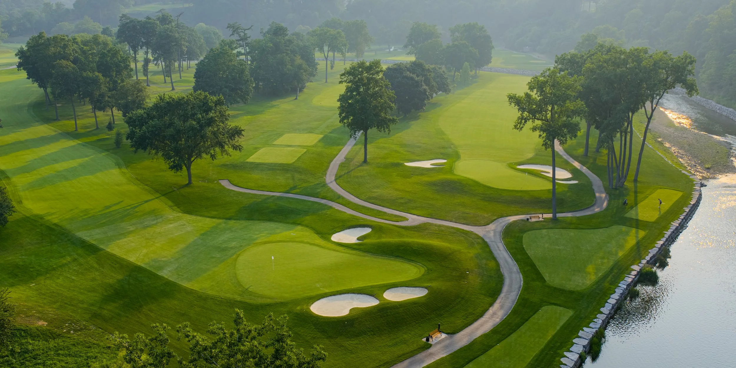 A scenic view of a golf course with green fairways, trees, sand traps, and a water hazard, viewed from above.